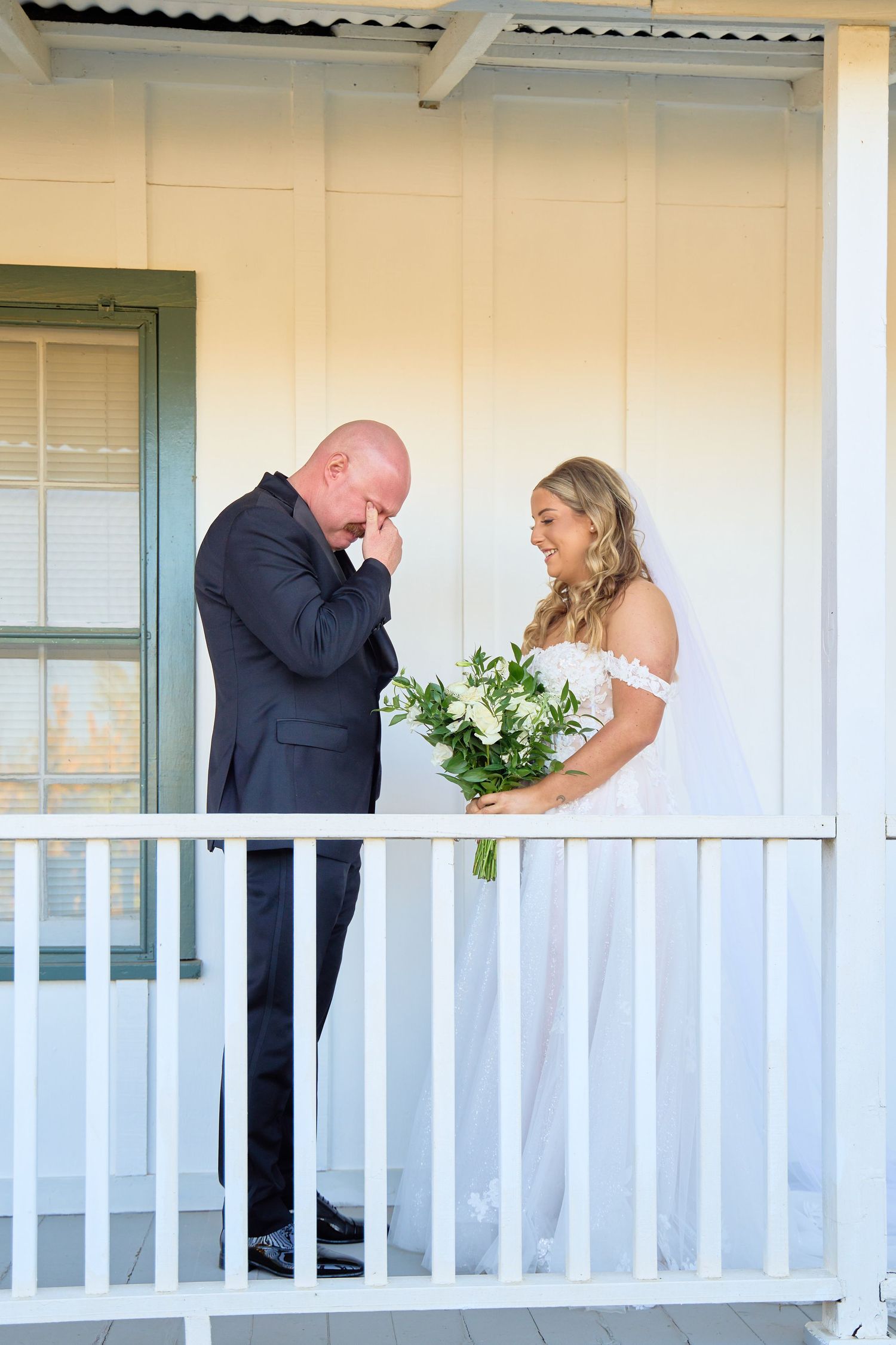 Groom emotional at first look with smiling bride holding bouquet on white porch.