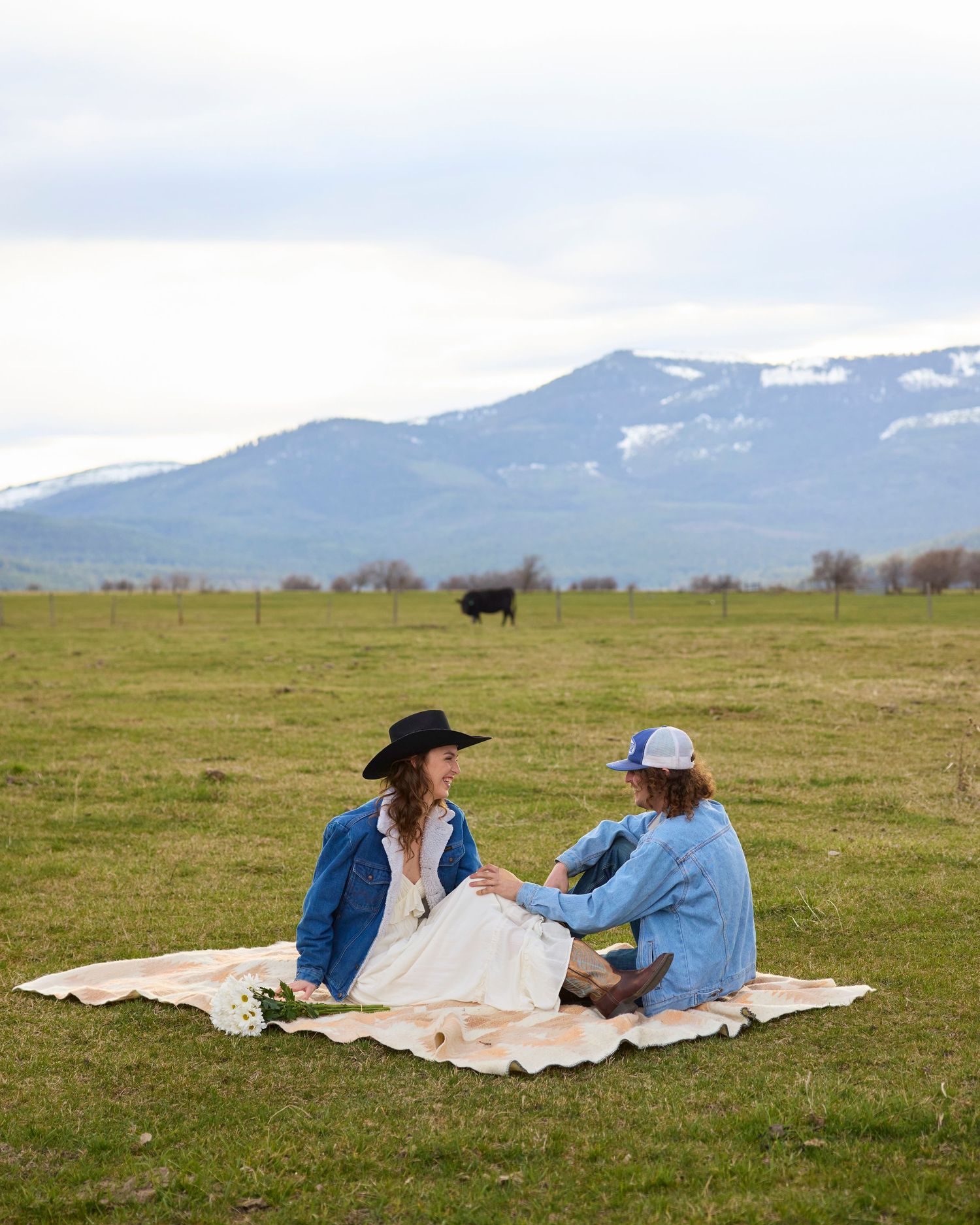 Couple sitting on blanket in green field with mountain backdrop and cow, bride in white dress and black hat.