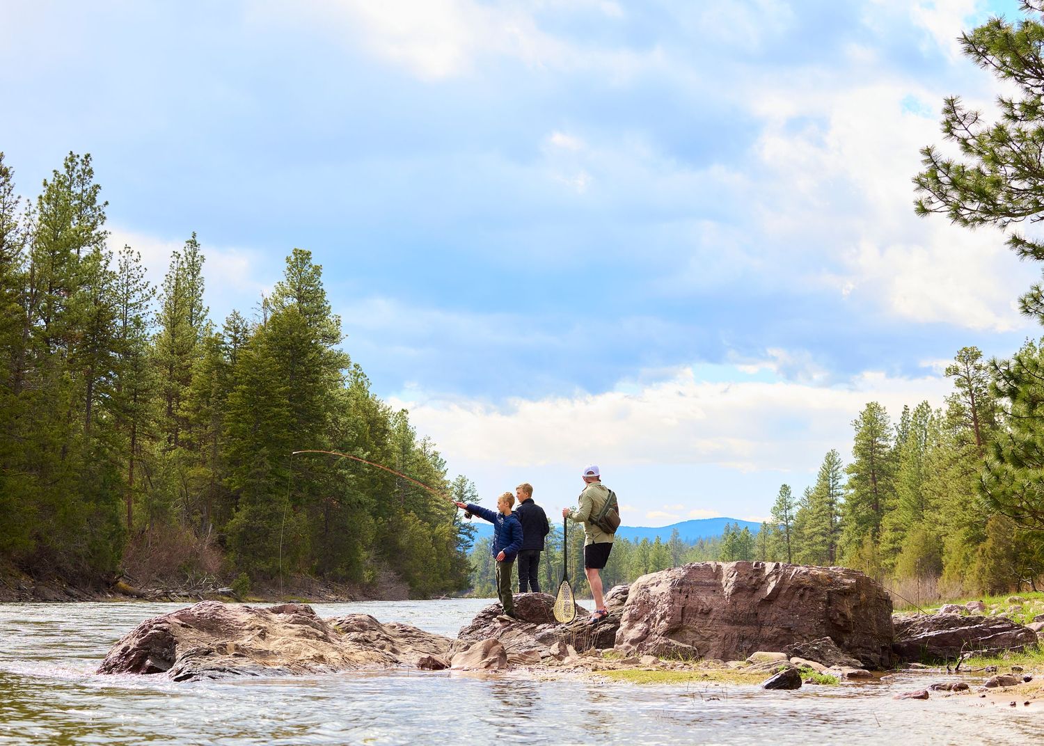 Three people standing on rocky riverbank pointing at scenic forested mountain landscape under cloudy sky.