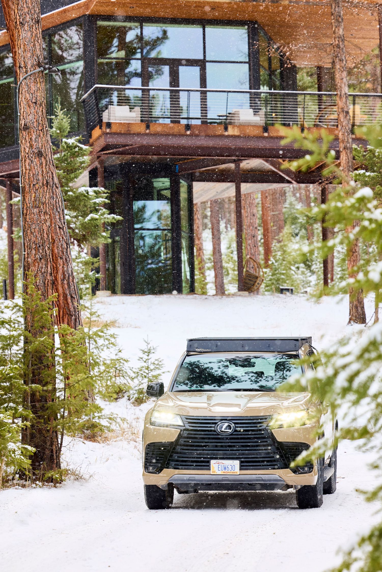 Gold Lexus LX SUV driving through snowy forest path toward modern luxury cabin.