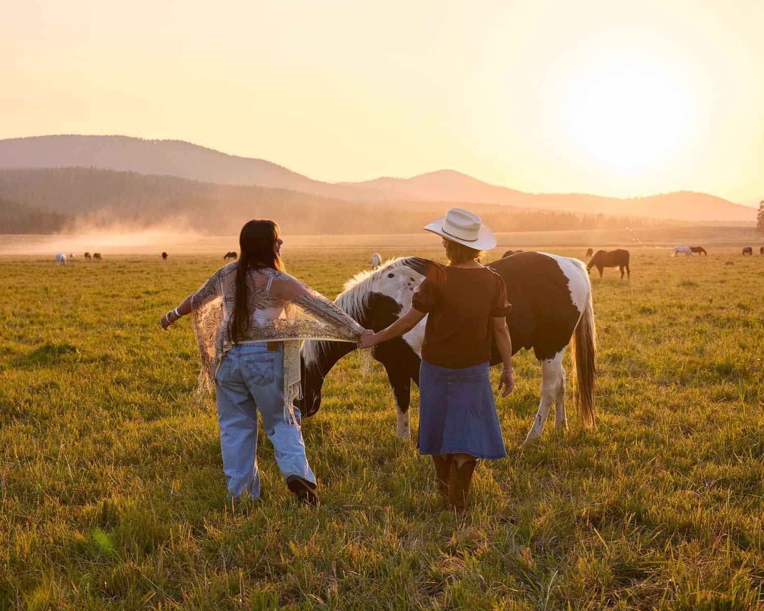 Two women walking with a paint horse through a misty golden sunrise meadow.