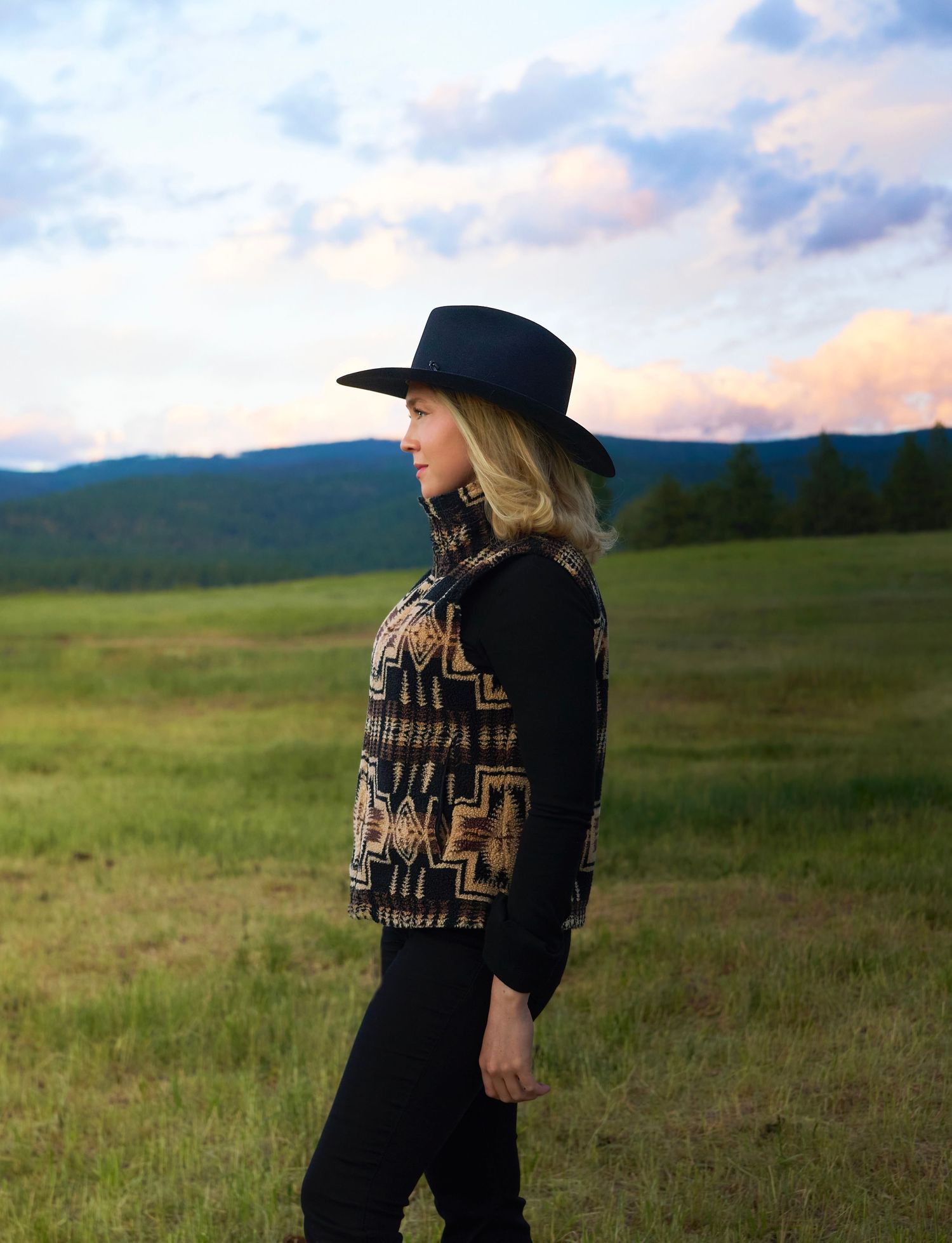 Blonde woman in black cowboy hat and Aztec-print jacket standing in a green meadow at sunset.