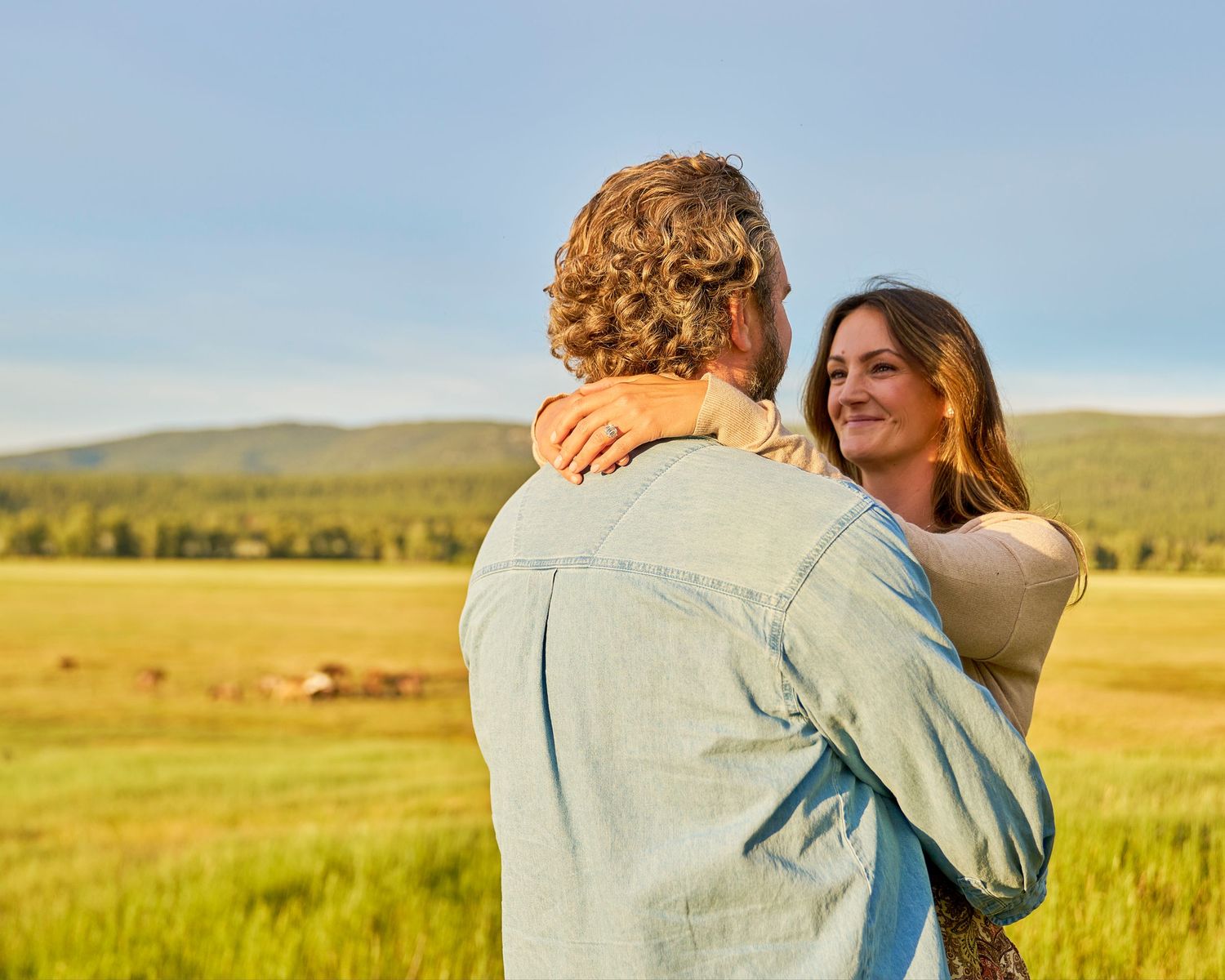 Woman smiling while embracing a man in a sunlit green field with rolling hills and farmland in the background.