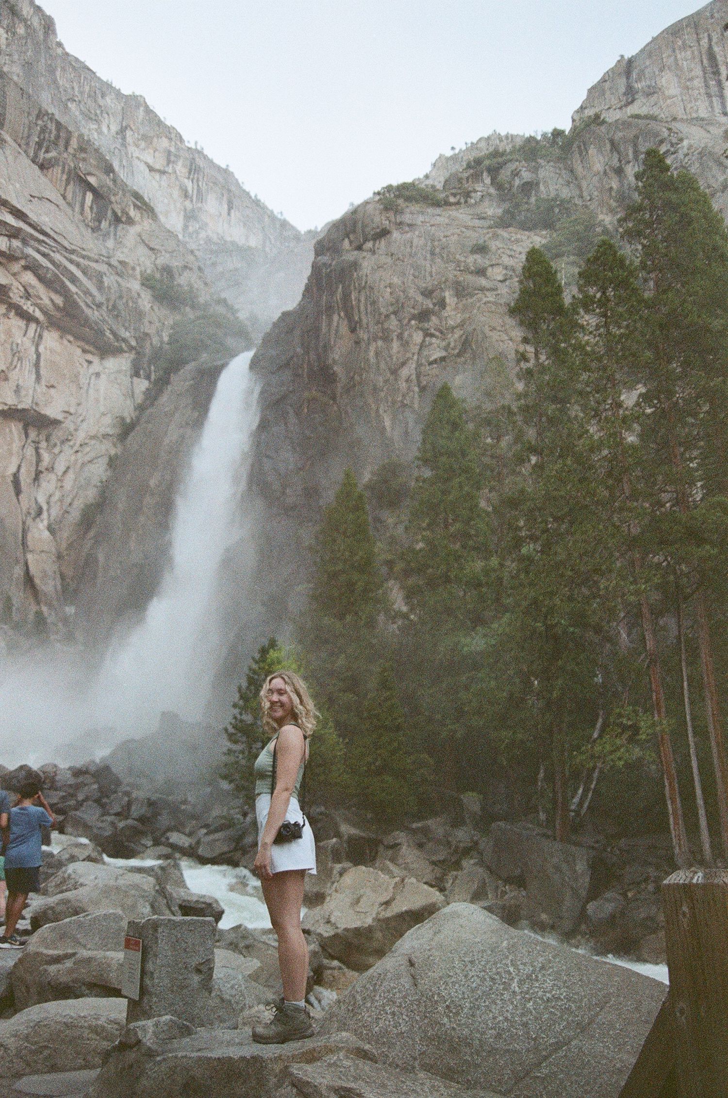 Blonde woman standing on rocks before massive Yosemite waterfall cascading down granite cliffs.