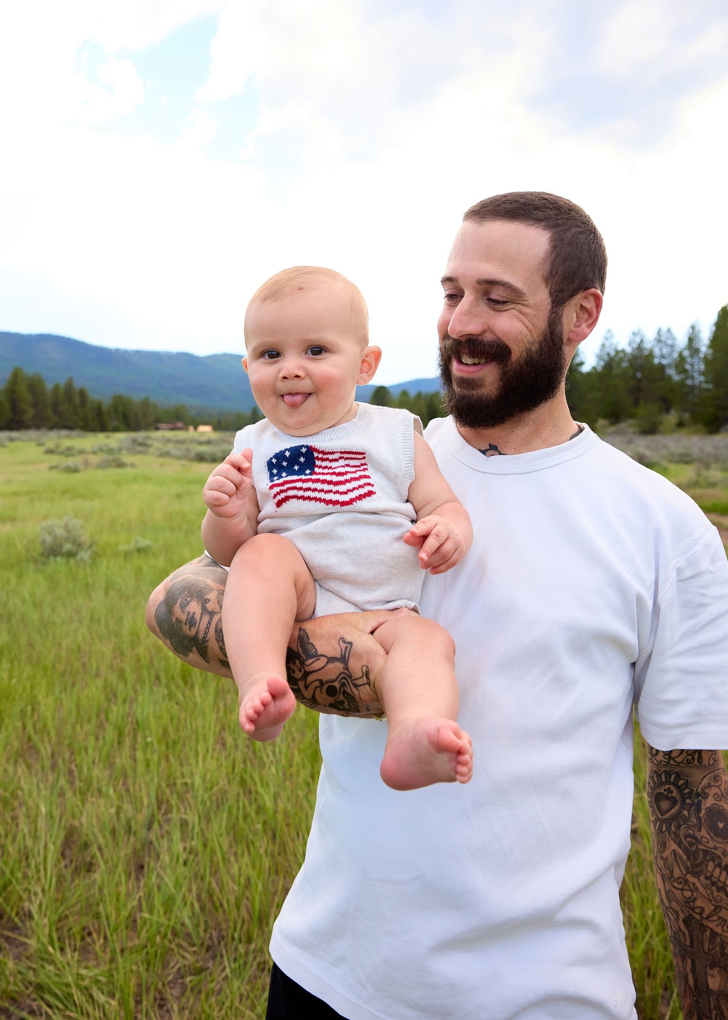 Smiling bearded man with tattoos holding a happy baby wearing an American flag onesie outdoors.