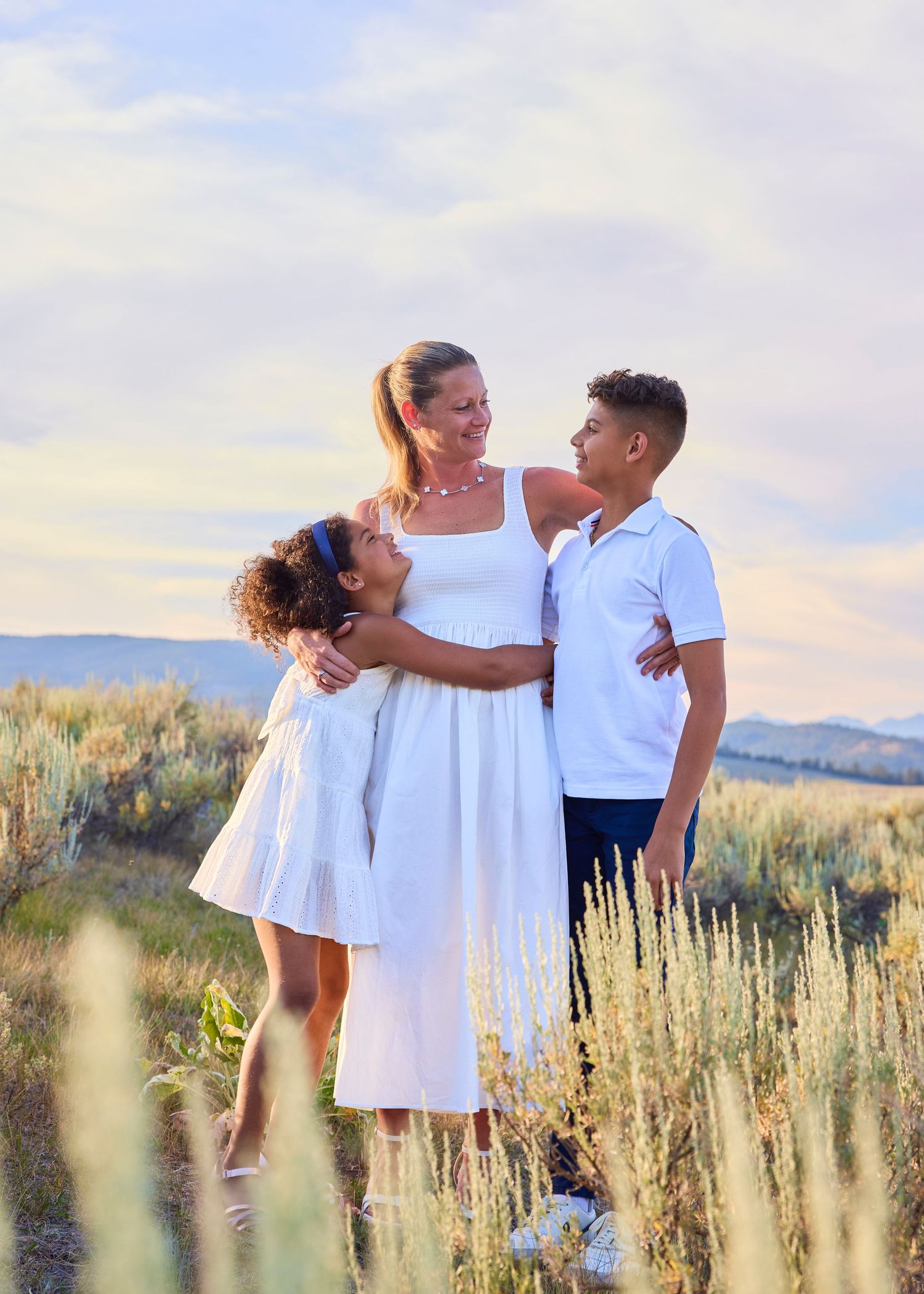 A mother in a white dress shares a warm embrace with her two children in a sagebrush meadow.