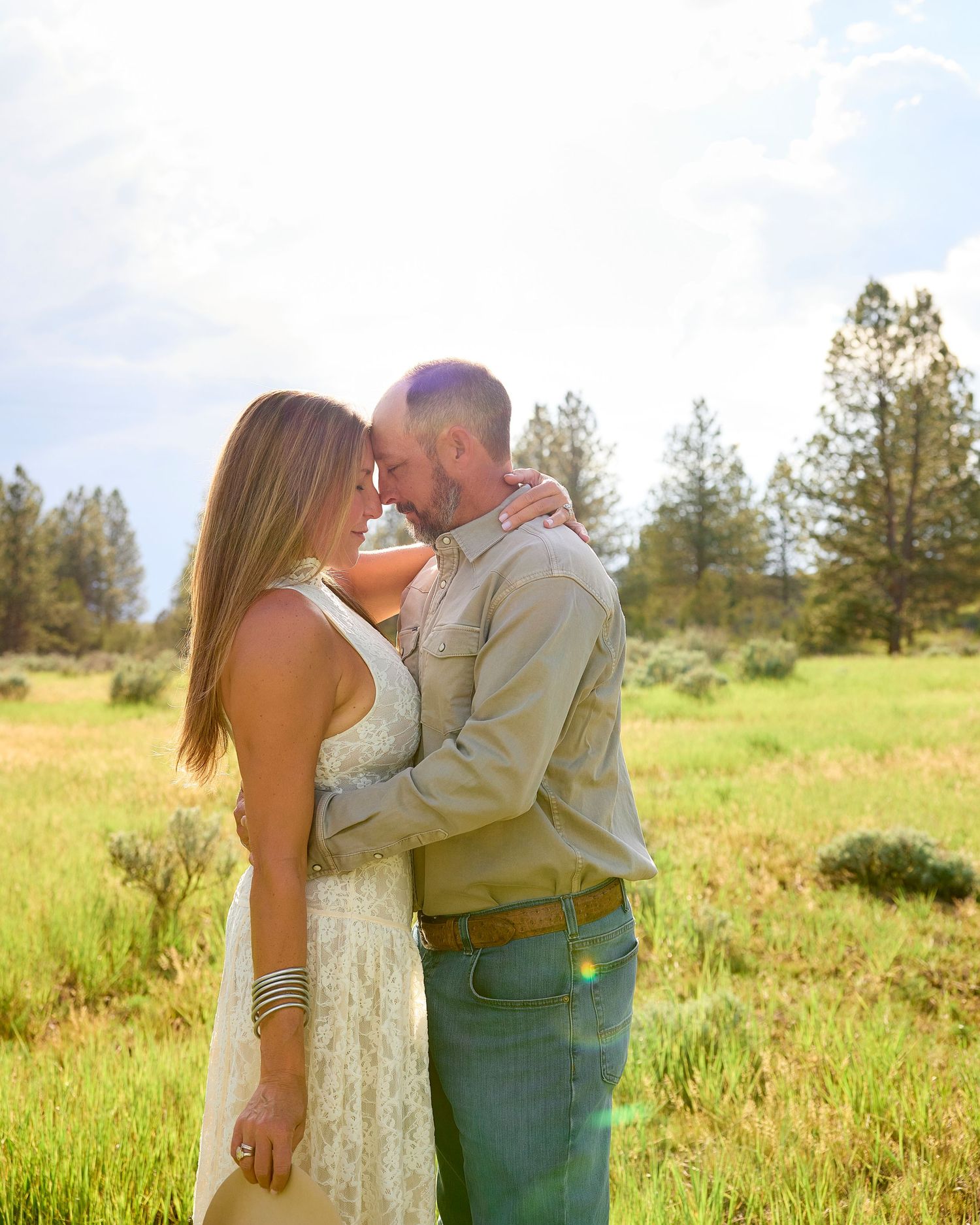 Romantic couple embracing forehead-to-forehead in a sunny green meadow with pine trees behind them.
