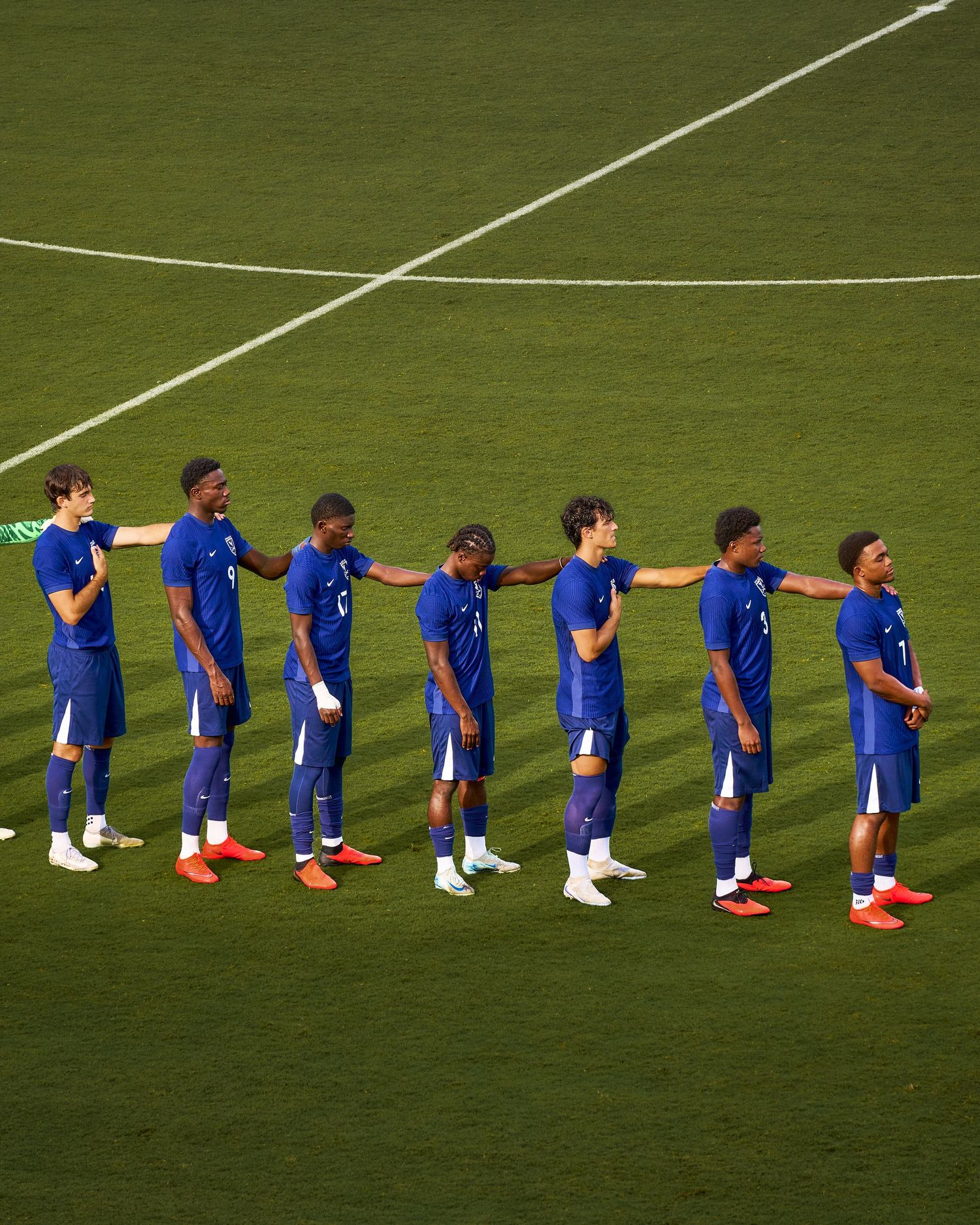 Seven soccer players in blue uniforms stand in a line on a green field during a pre-match ceremony.