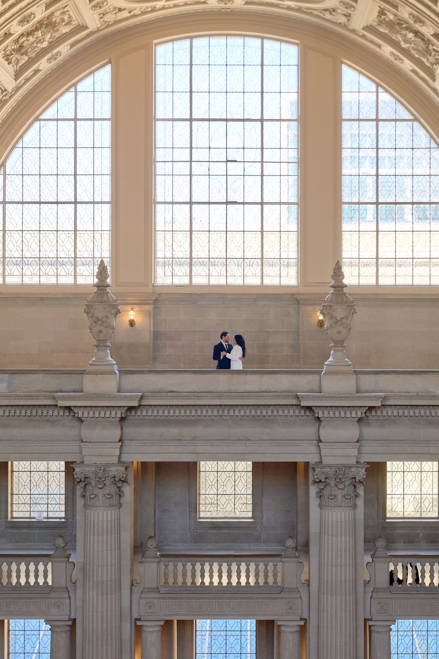 Couple embraces on ornate balcony of San Francisco City Hall with grand arched windows.