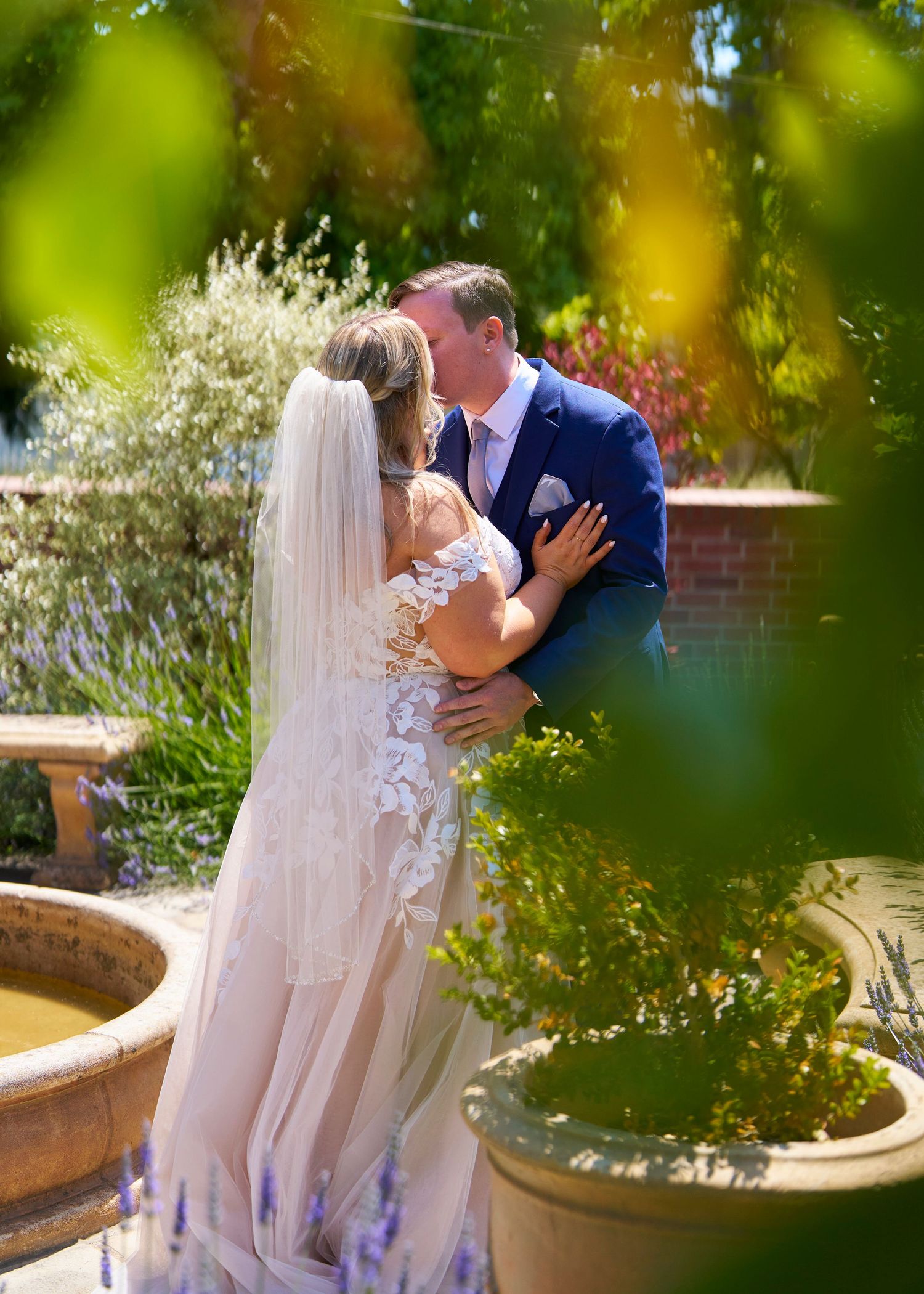 Bride and groom sharing a romantic kiss in a lush garden, groom wearing blue suit, bride in lace gown.