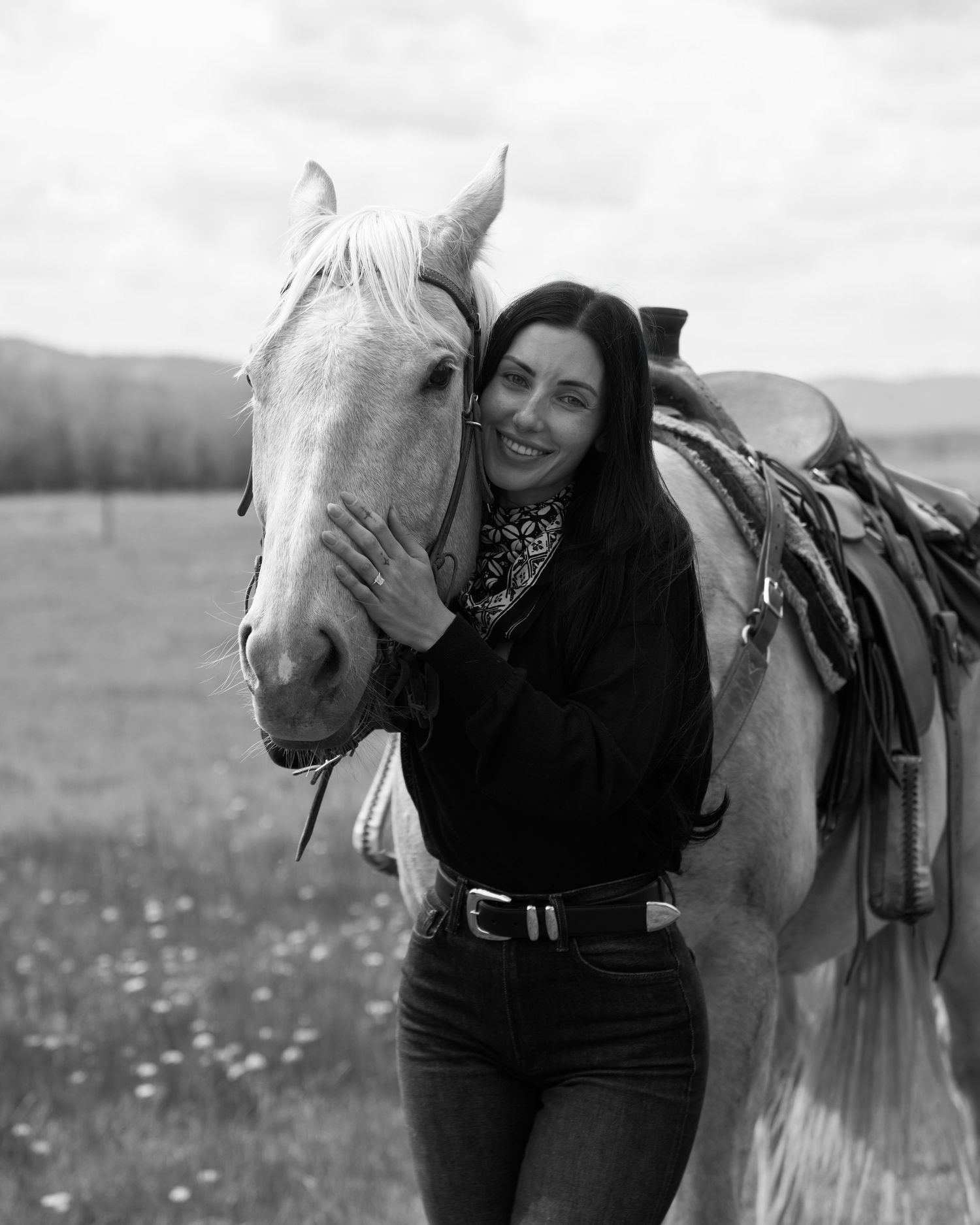 Black and white portrait of smiling woman embracing saddled palomino horse in field.