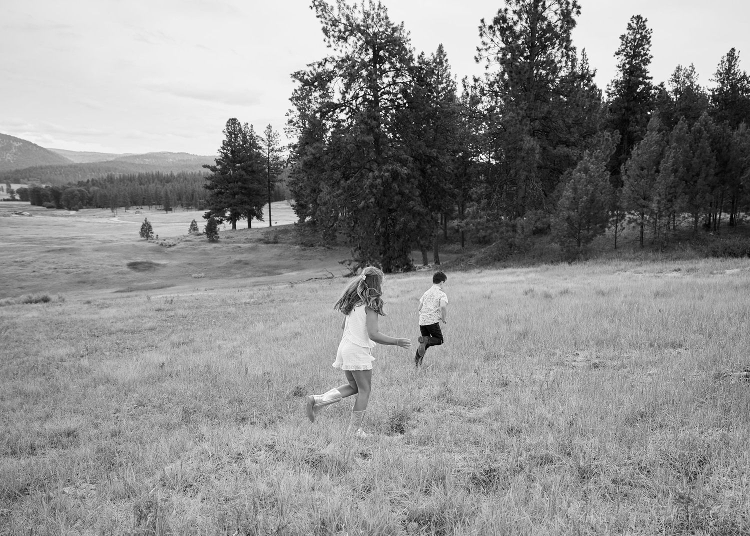Black and white photo of two children running through an open meadow surrounded by pine trees.