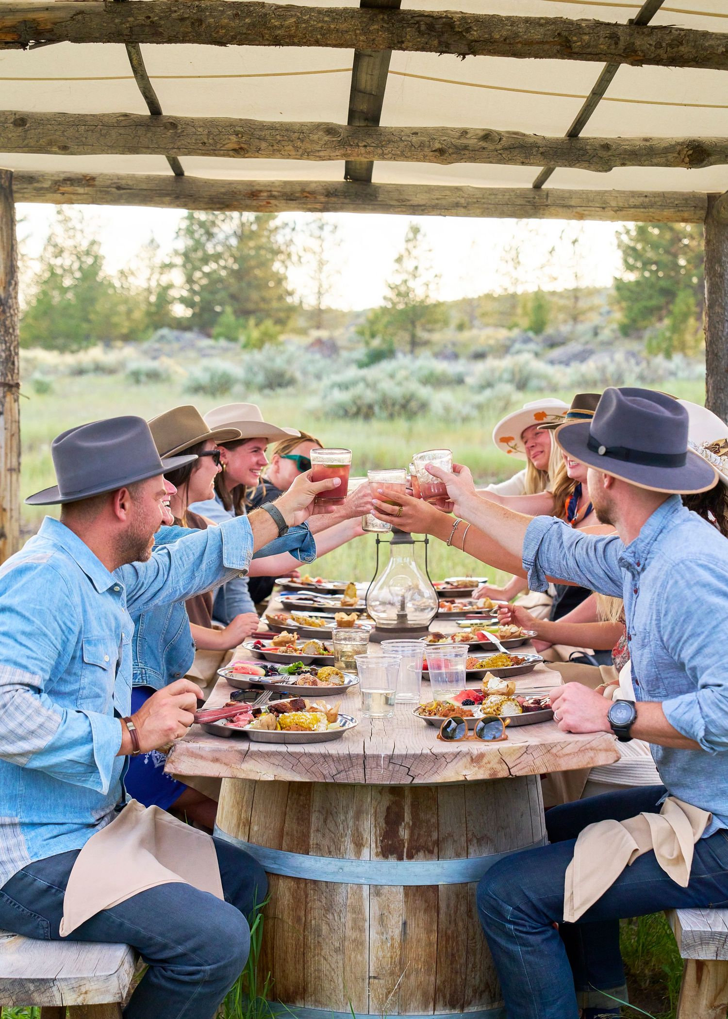 Group of friends in cowboy hats toast drinks at a rustic outdoor ranch dining table.