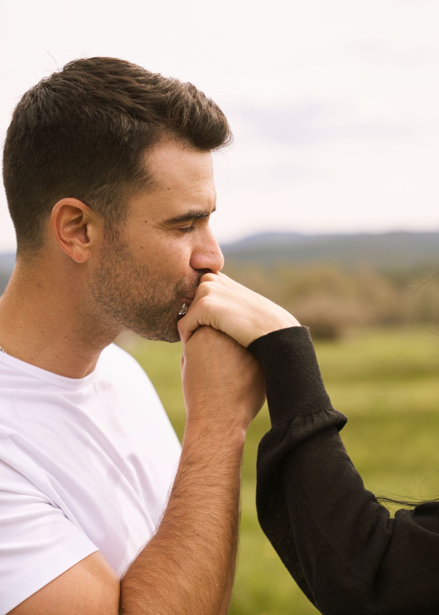 Man in white t-shirt gently kissing a woman's hand outdoors with a blurred green landscape in the background.