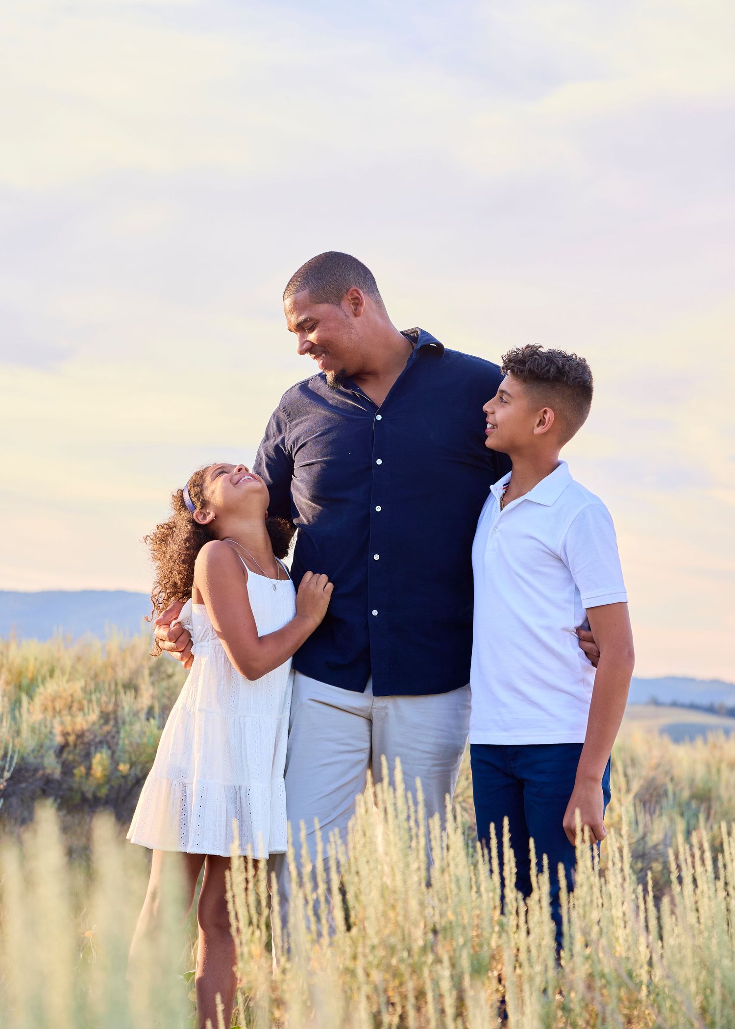 A tall father smiles warmly while embracing his two children in a golden sagebrush field at sunset.