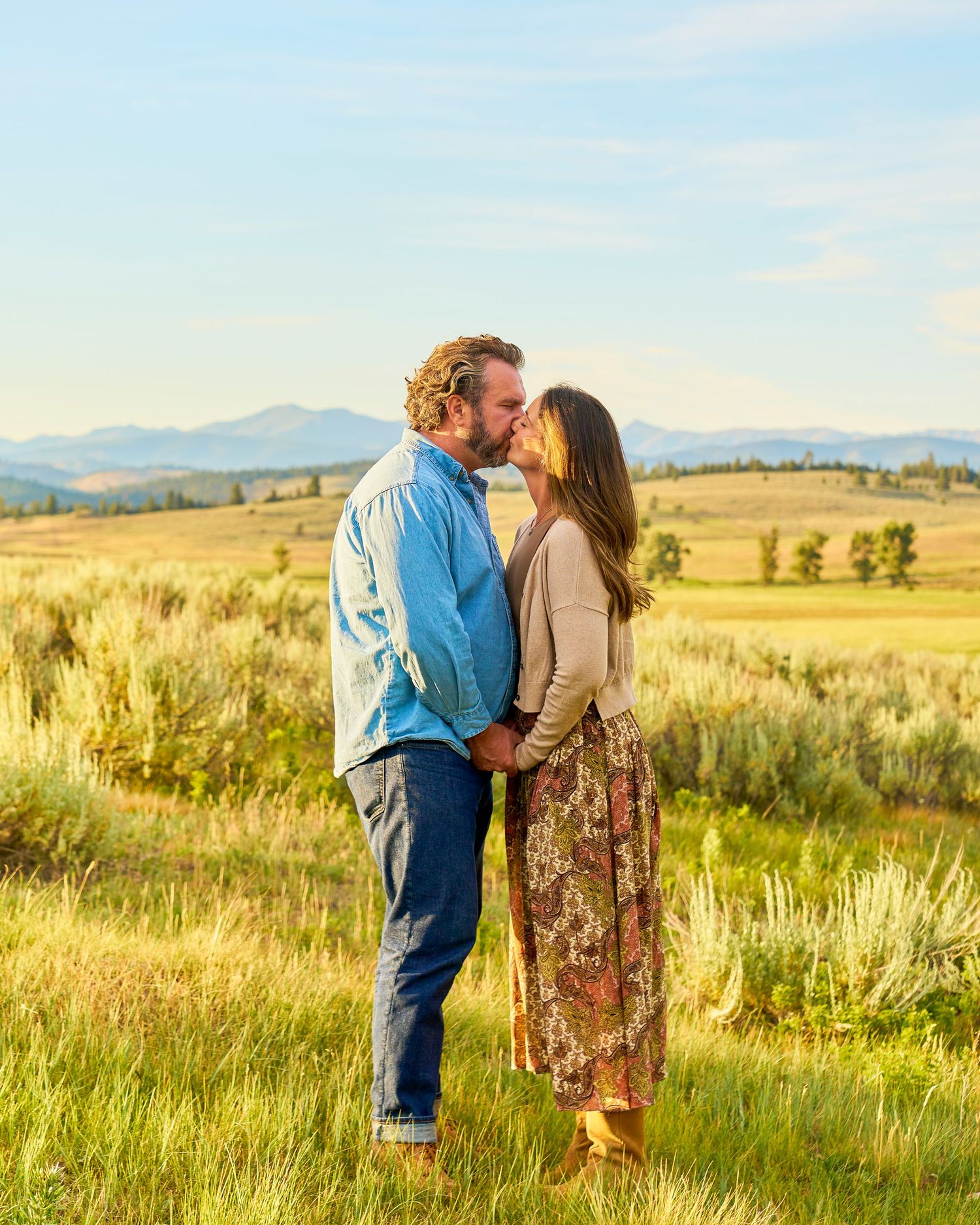 Couple shares a kiss in golden sagebrush field with distant mountain range under blue sky.