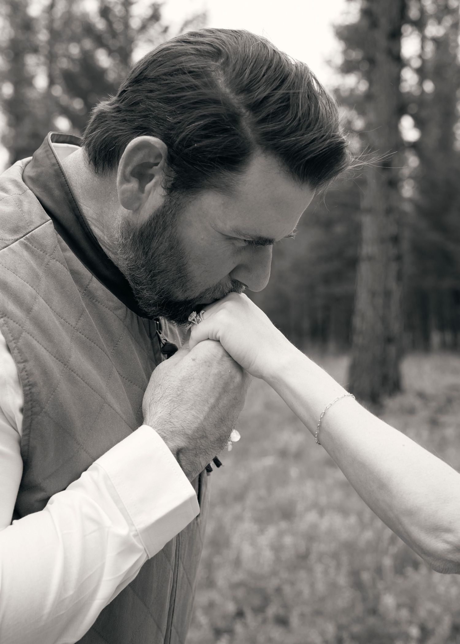 Black and white photo of a bearded groom in a vest tenderly kissing the bride's hand in a pine forest.