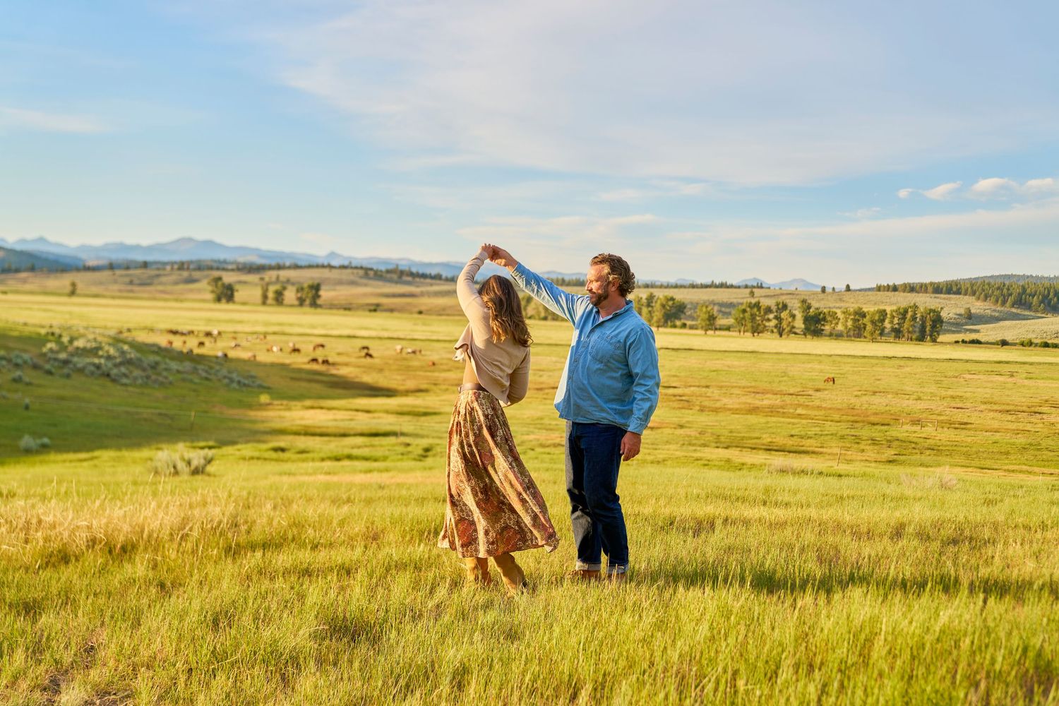 Romantic couple dancing together in a sunlit green field with scenic mountain landscape and grazing livestock behind them.