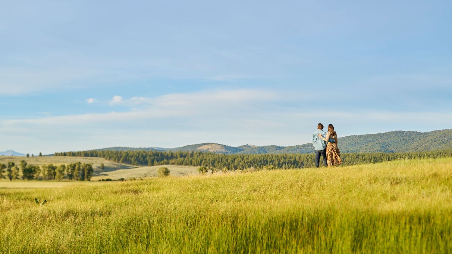Couple stands together in expansive golden grass meadow with forested hills in the distance.