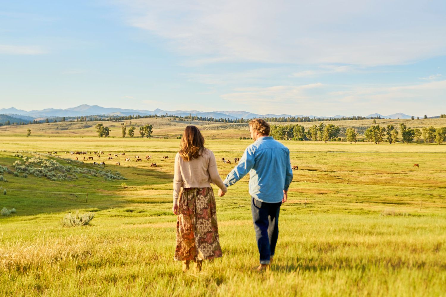 Couple holds hands walking through golden meadow with vast valley and mountain views behind them.