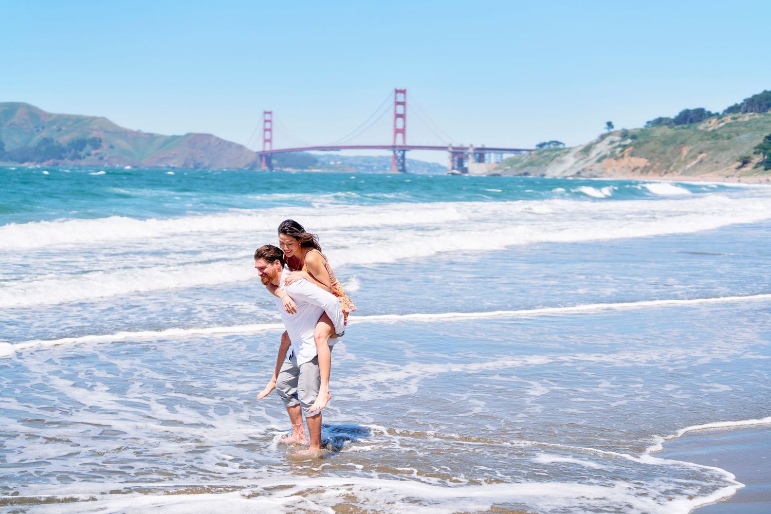 Couple playing in ocean waves at Baker Beach with Golden Gate Bridge in background.