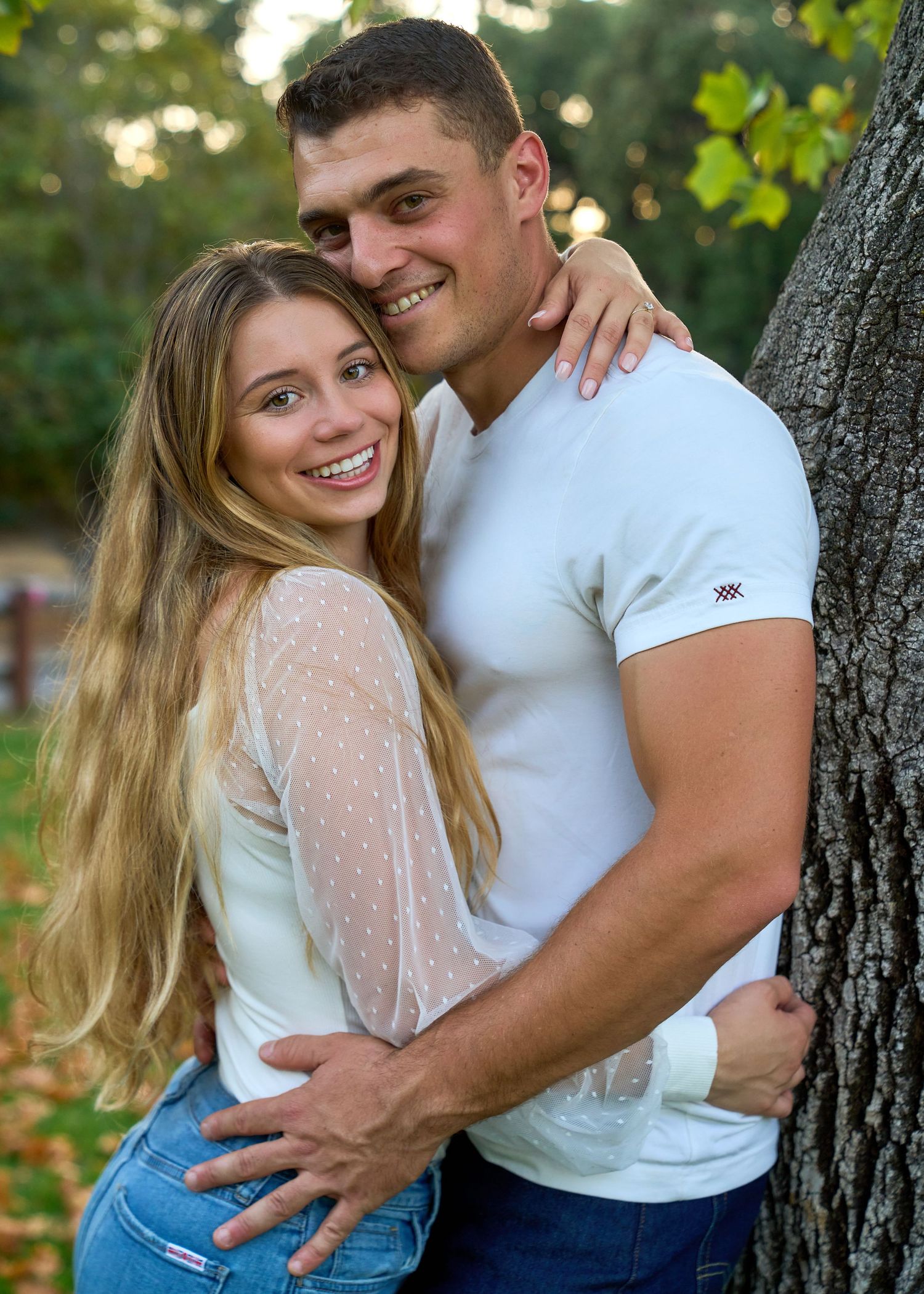 Happy young couple embracing by tree in park during golden hour engagement photo session.