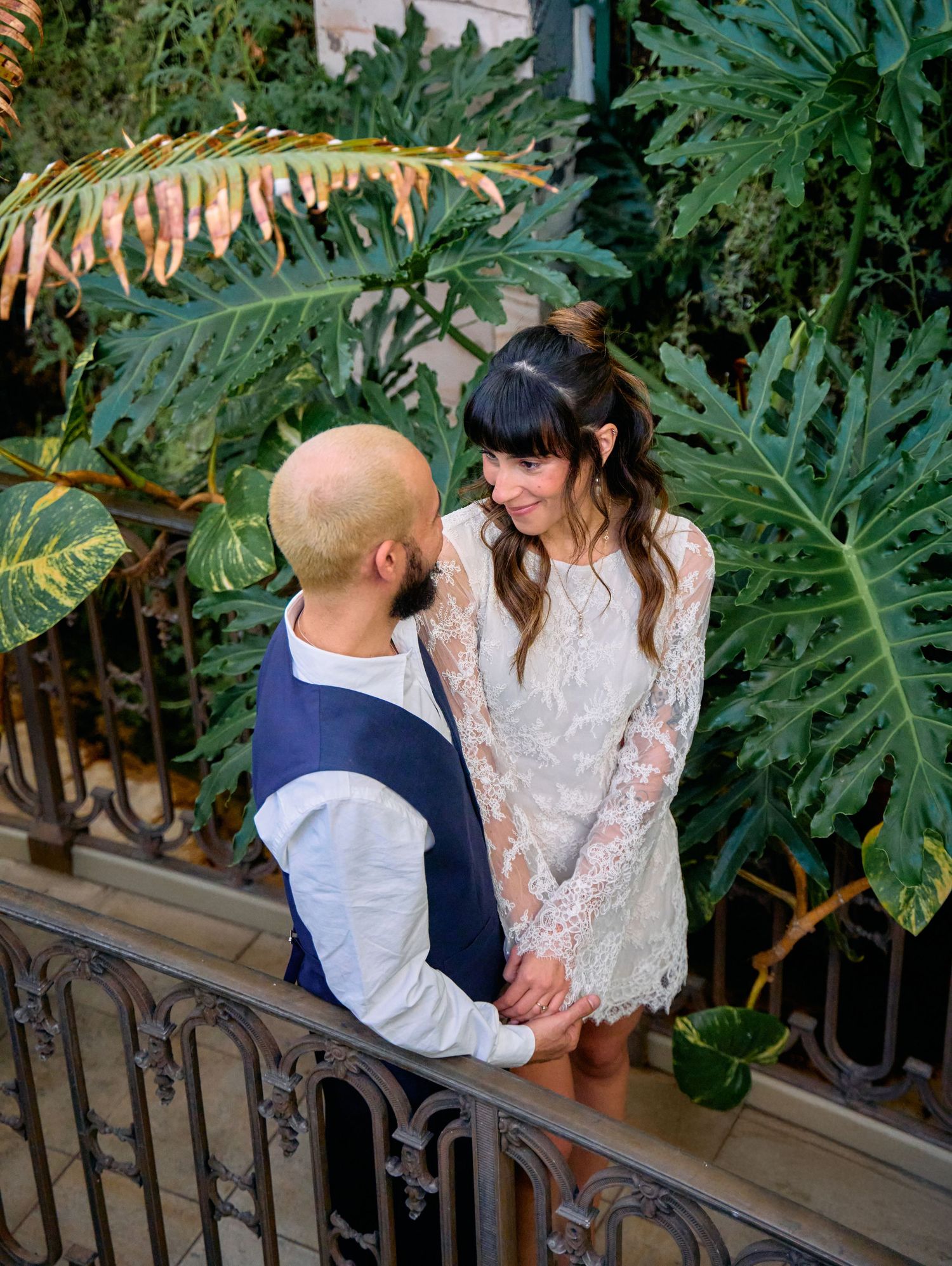 Newlyweds hold hands and gaze at each other surrounded by lush tropical plants on a balcony.