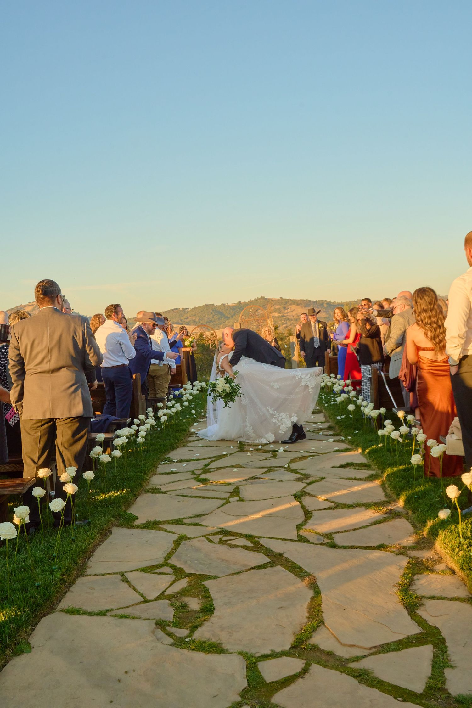 Bride and groom share romantic dip kiss at outdoor sunset wedding ceremony.