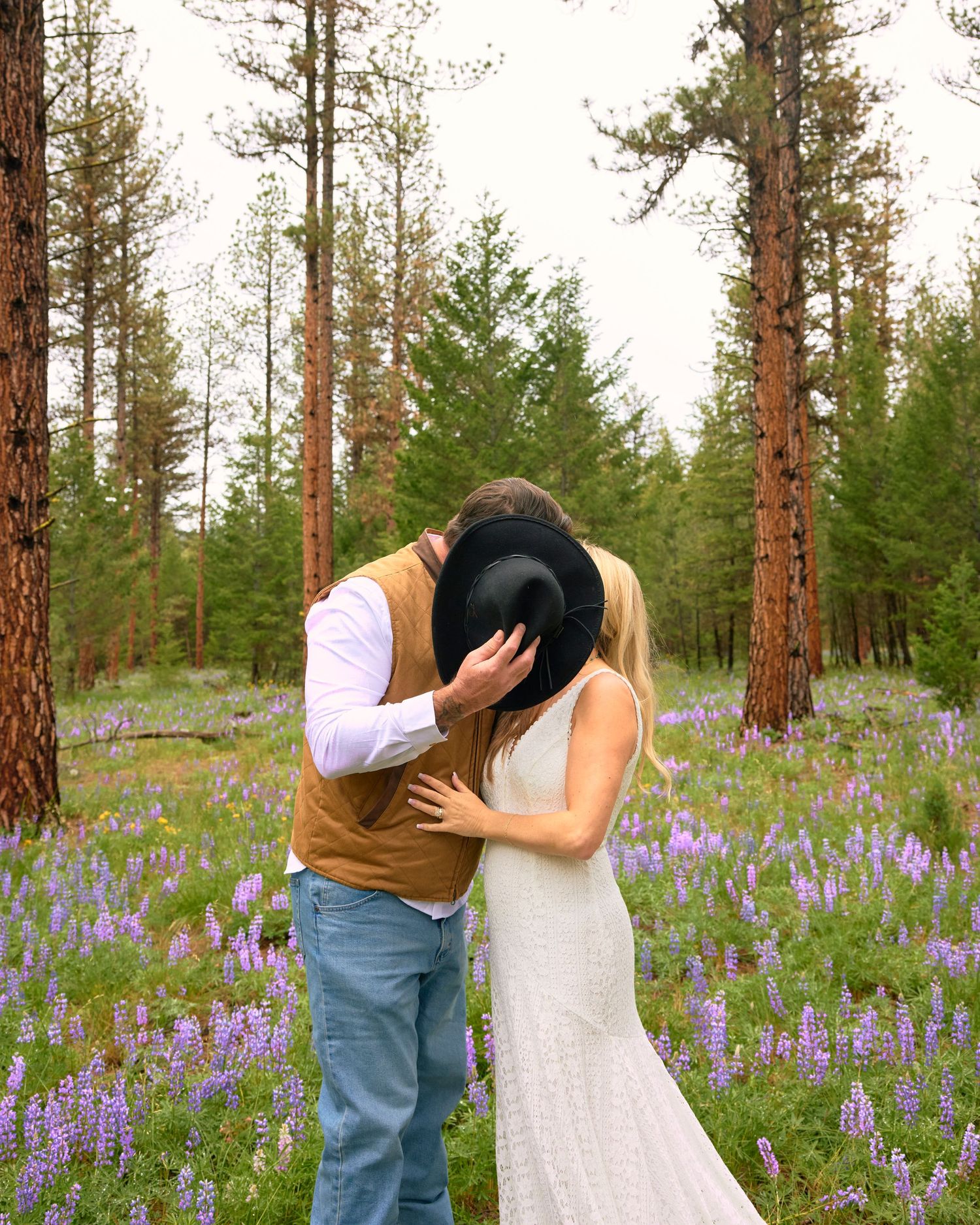 Couple sharing a romantic kiss in a wildflower meadow surrounded by tall pine trees, groom hiding behind black cowboy hat.