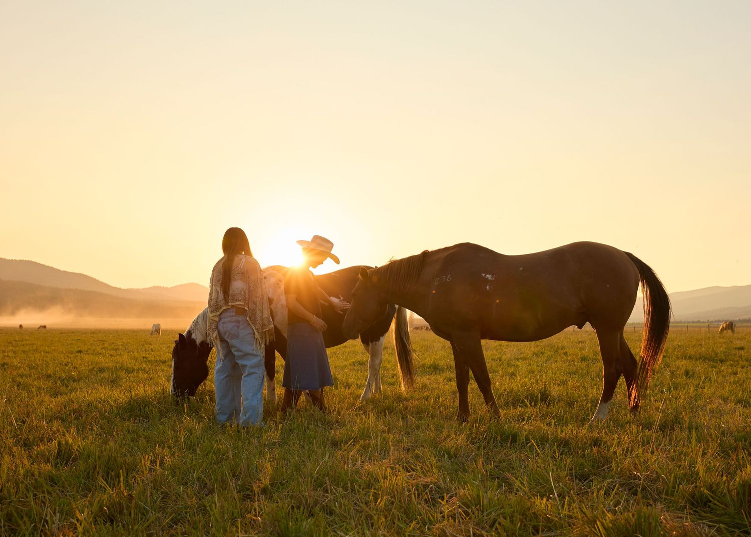 Two women stand with horses in a golden sunset field with misty mountains in the background.