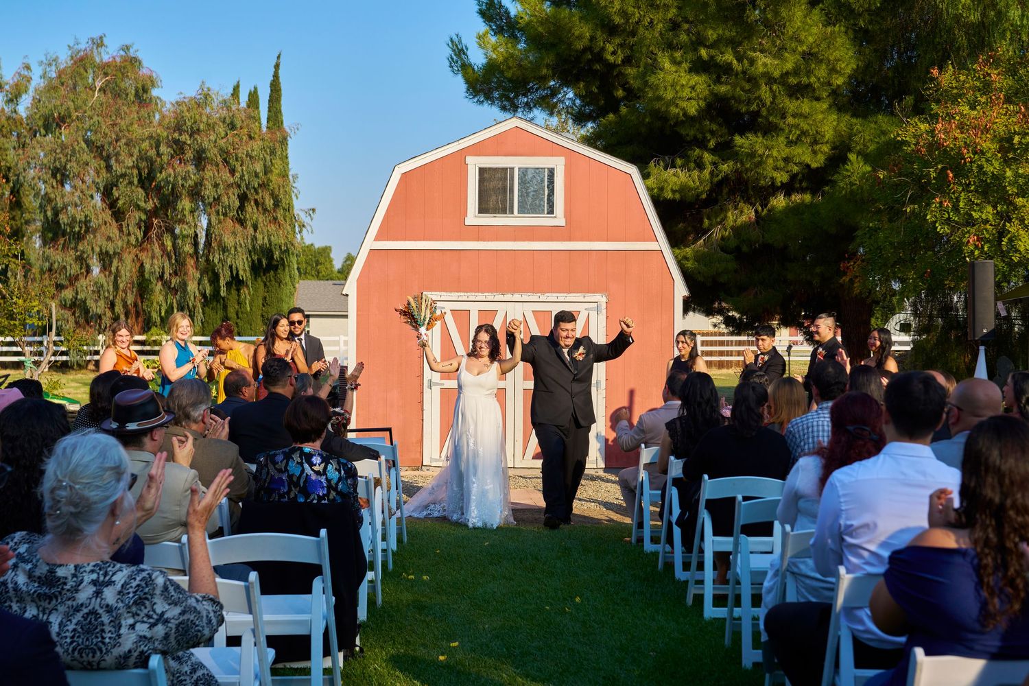 Outdoor wedding ceremony on green lawn with guests seated in white chairs facing couple before a red barn.