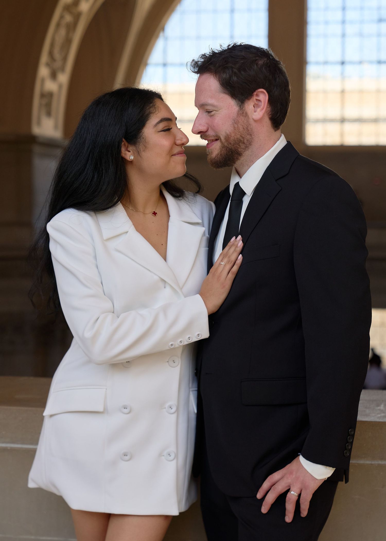 Couple shares romantic nose-to-nose moment, bride in white blazer dress touching groom's tie indoors.