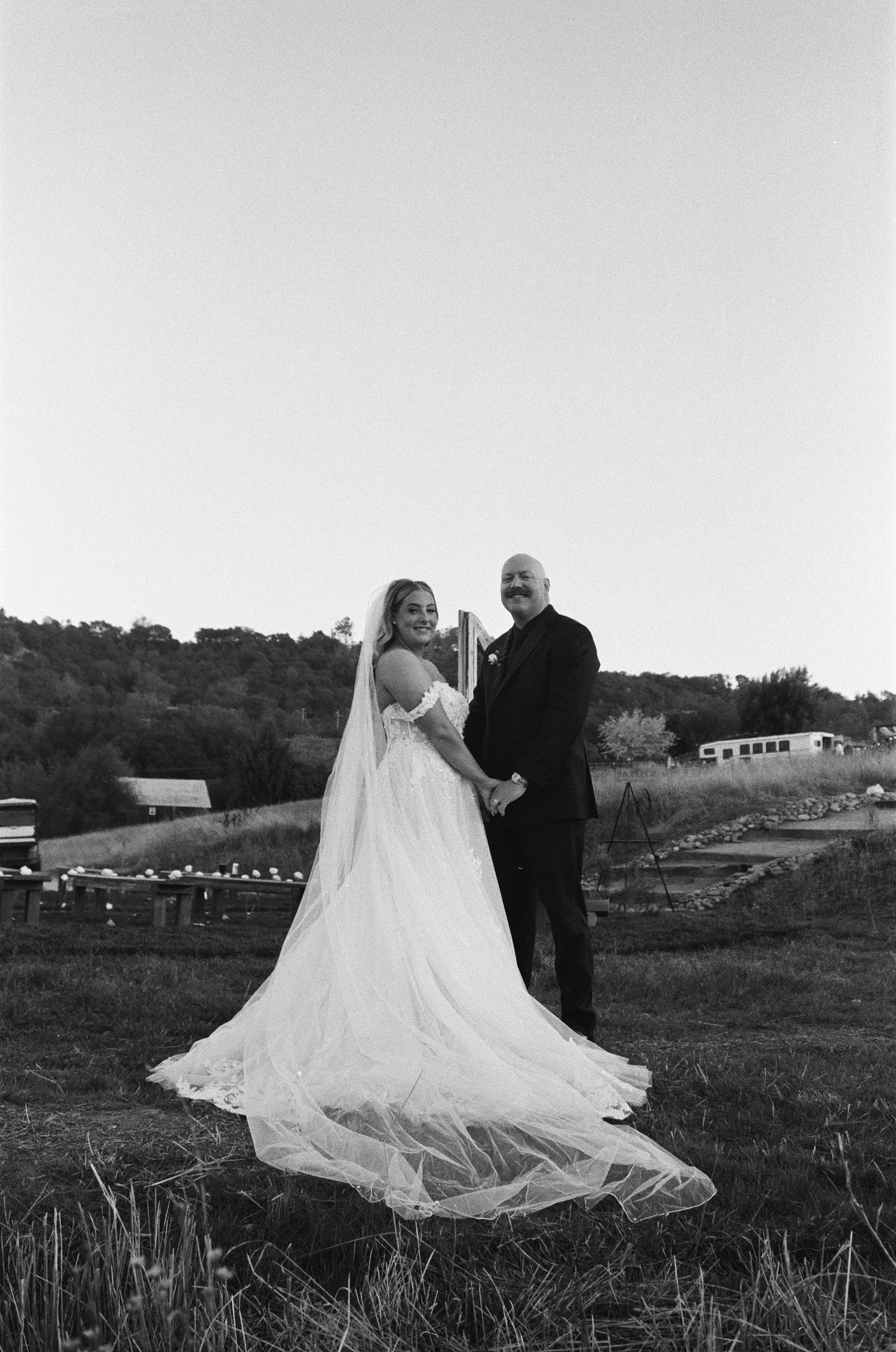 Black and white portrait of bride in flowing gown and veil with groom holding hands in a scenic outdoor farm setting.