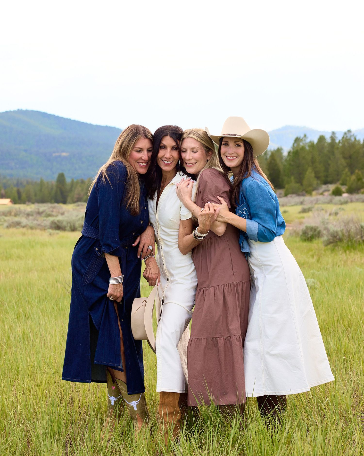 Four women hugging and smiling together in a green mountain meadow on a cloudy day.