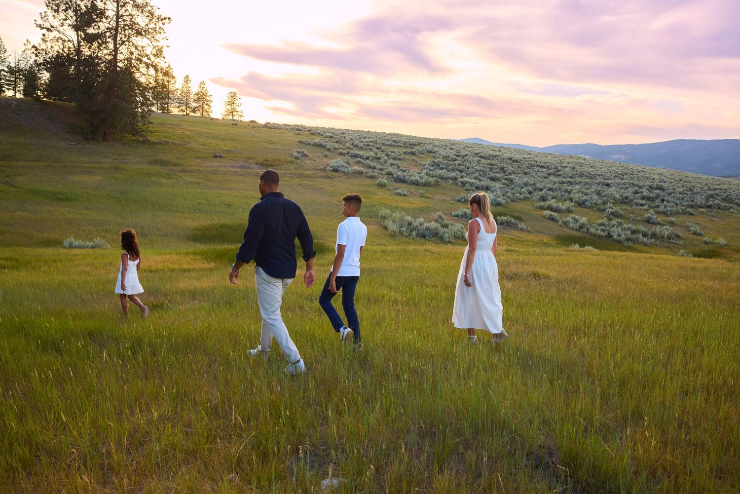 Family of four walks through a scenic green meadow at sunset with rolling hills and sagebrush landscape.