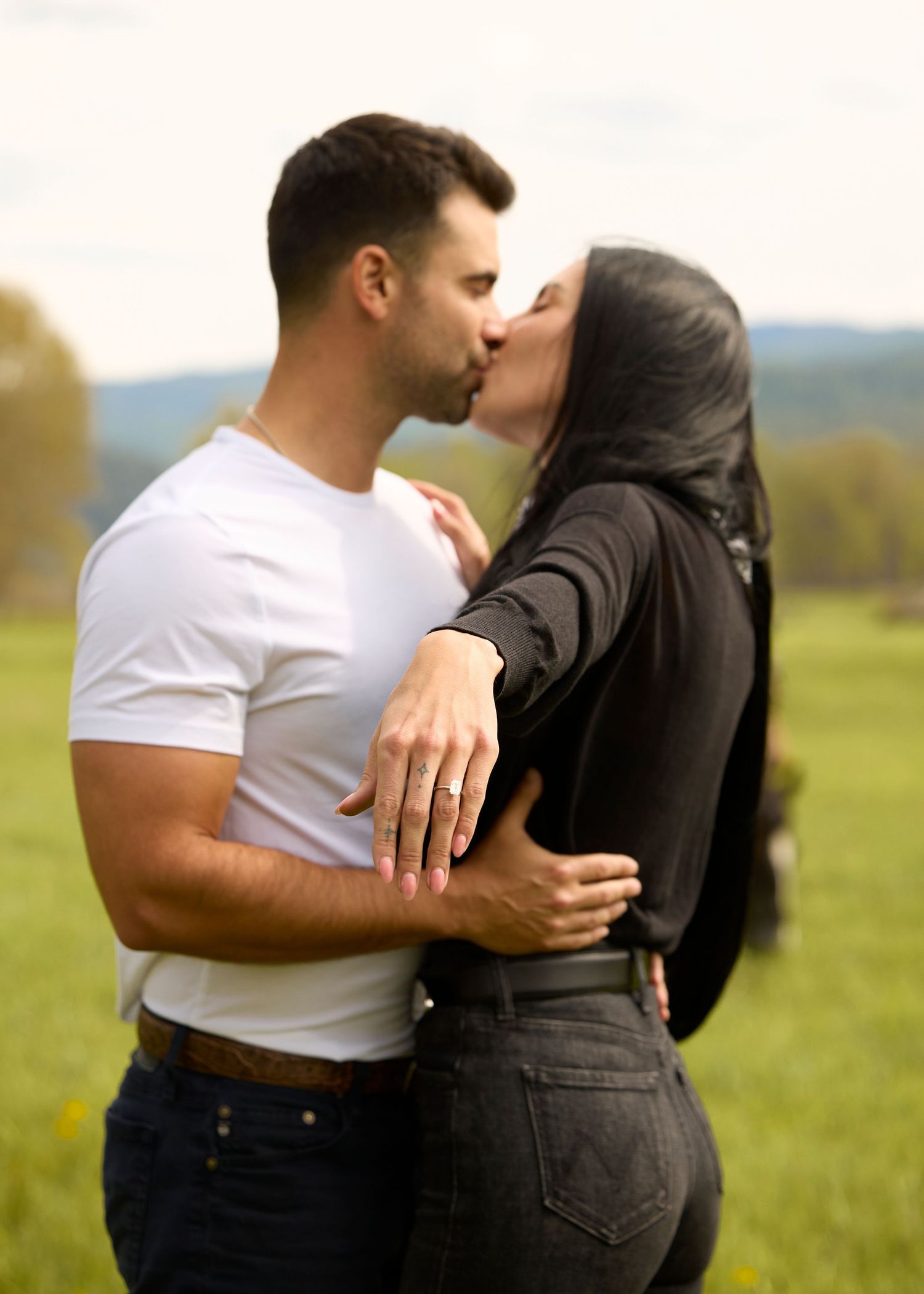 Couple sharing romantic kiss in lush green field, woman displaying engagement ring outdoors.