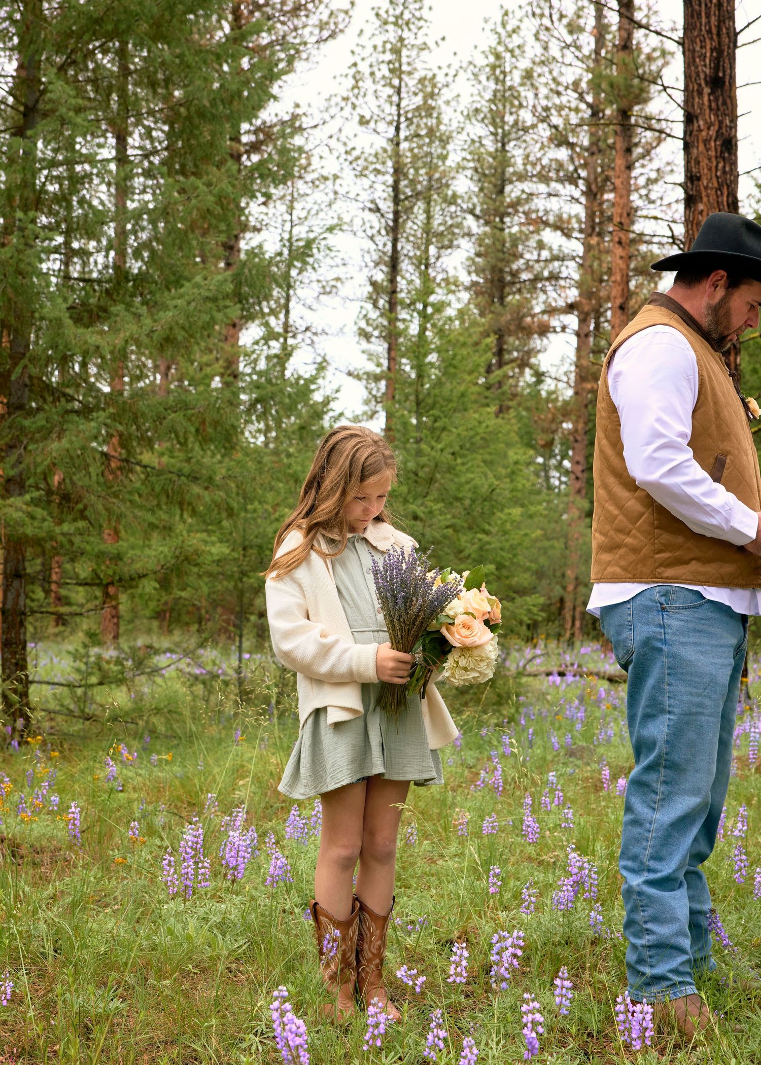 Young girl in green dress and cowboy boots admires a bouquet in a lupine meadow beside a man in a brown vest.