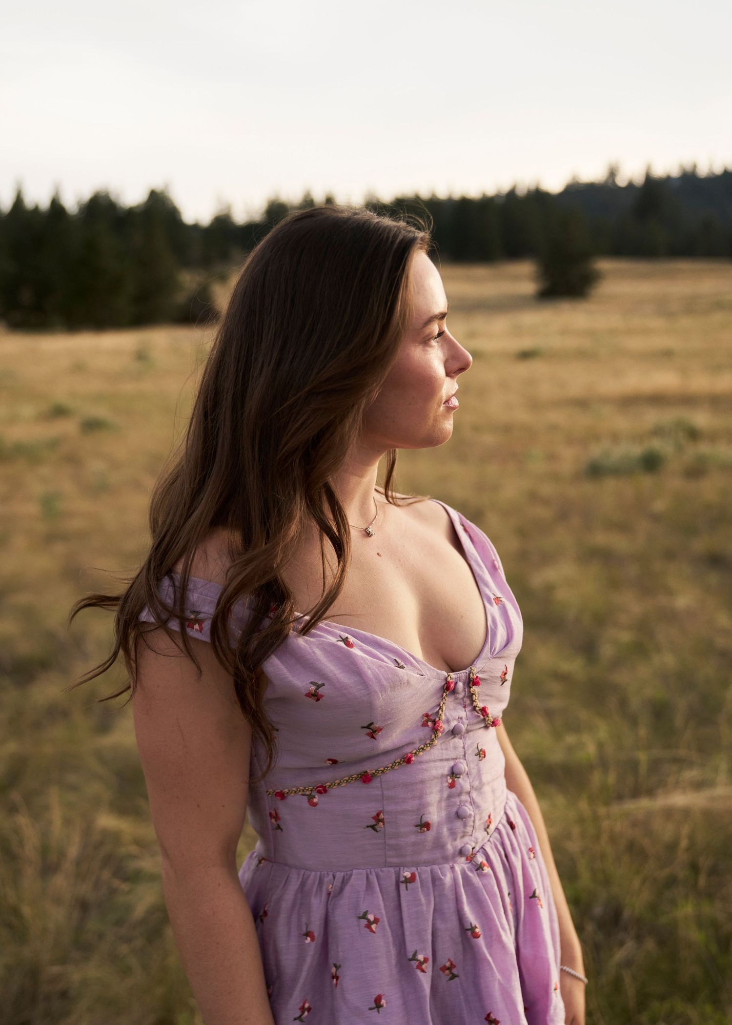 Woman in lavender floral dress stands in golden meadow with pine trees, looking sideways in warm sunset light.