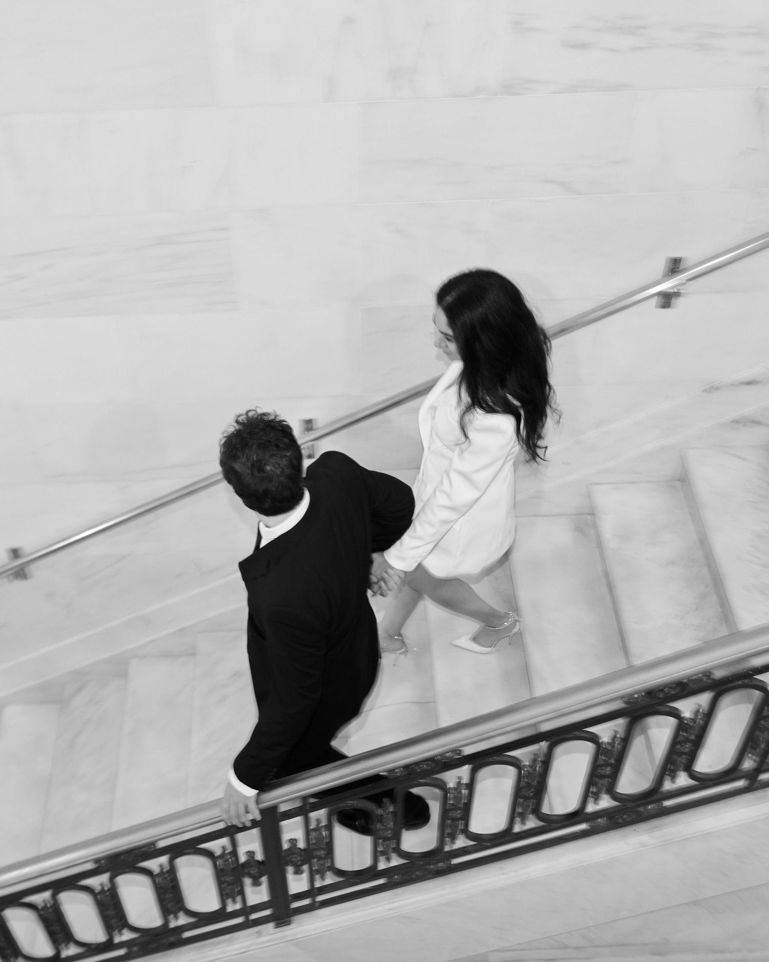 Aerial black and white photo of a couple walking up marble stairs together, woman in white blazer and man in black suit.