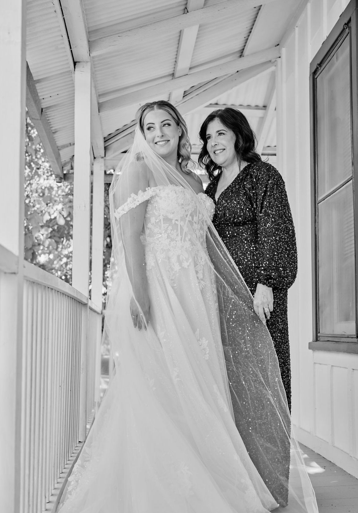 Bride in lace off-shoulder gown with veil poses with guest on wooden porch in black and white photo.