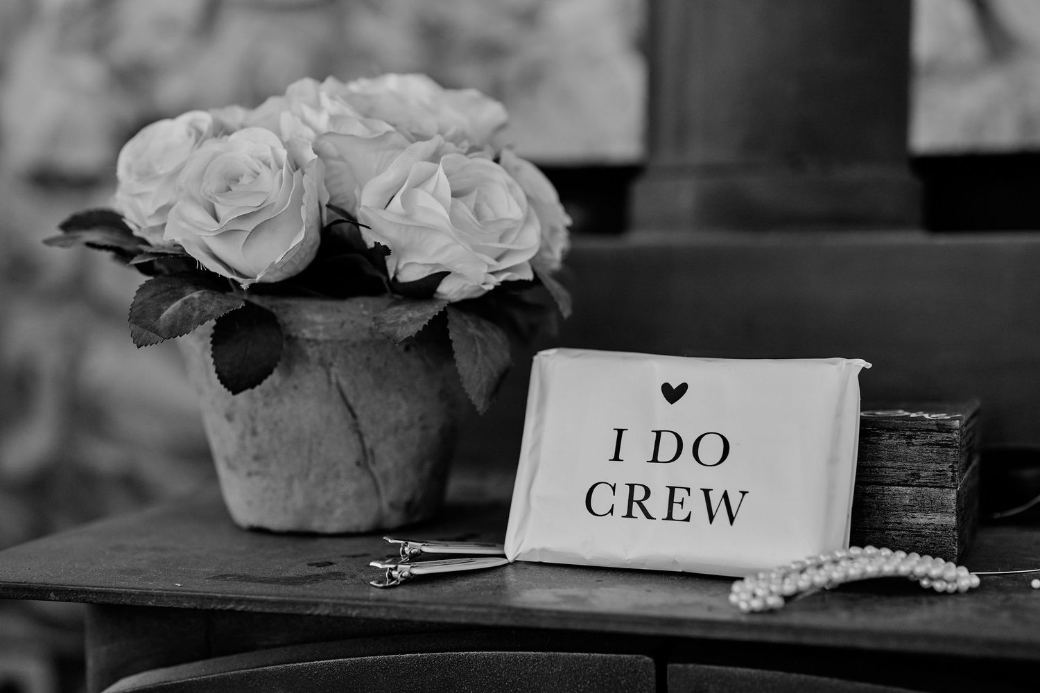 Black and white photo of roses in pot beside I Do Crew clutch and pearl necklace on table.