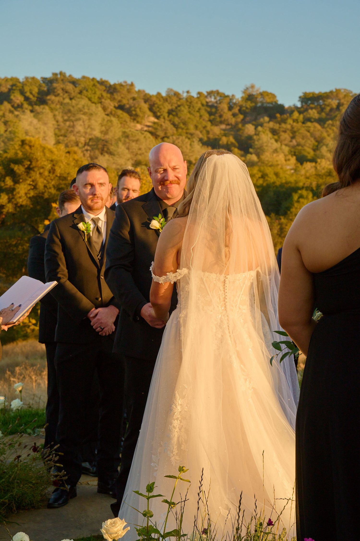 Groom smiles at bride during outdoor wedding ceremony at golden hour with rolling hills behind them.