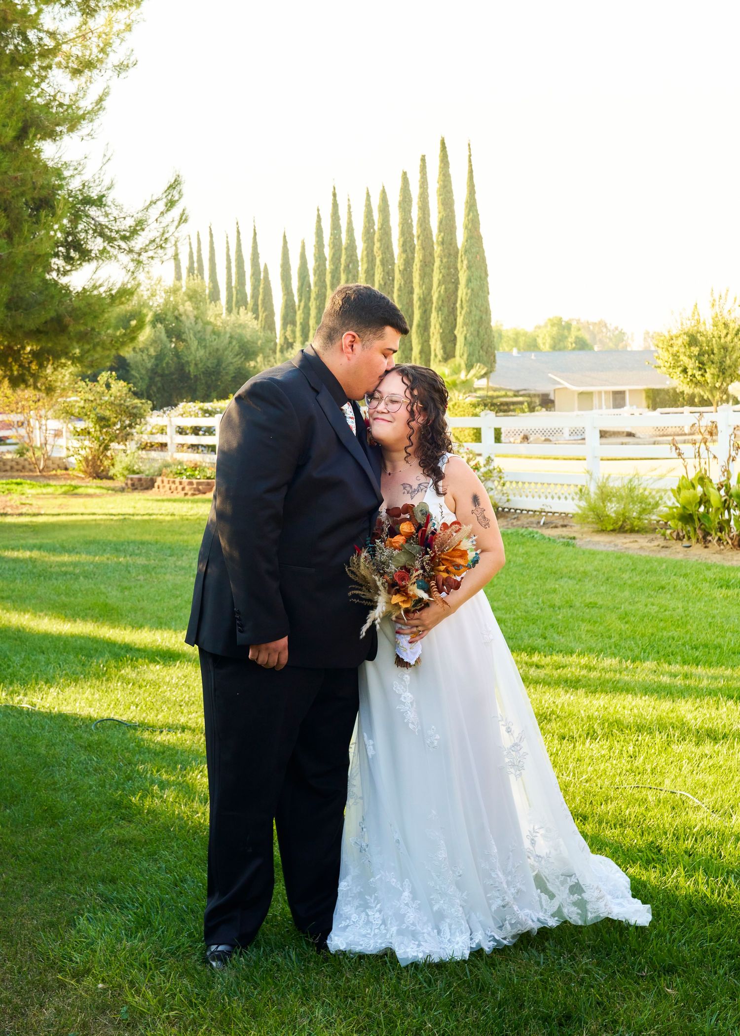 Groom kisses bride on cheek as she holds autumn bouquet on green lawn at sunset.