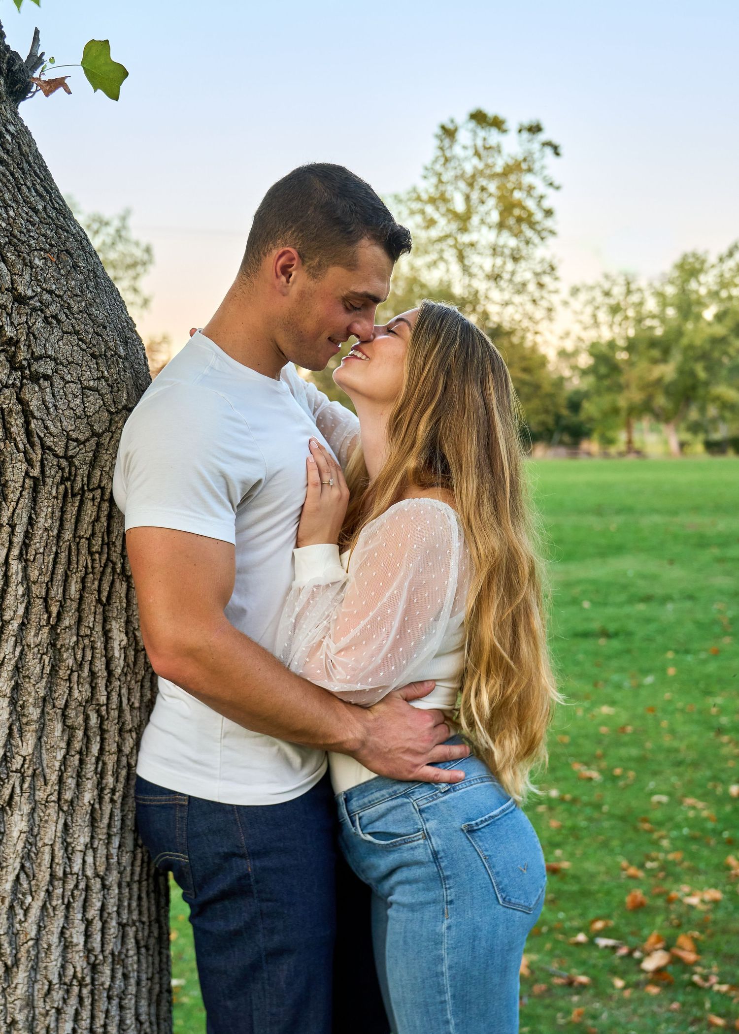 Happy couple sharing a kiss near a tree in a lush green park during engagement photos.