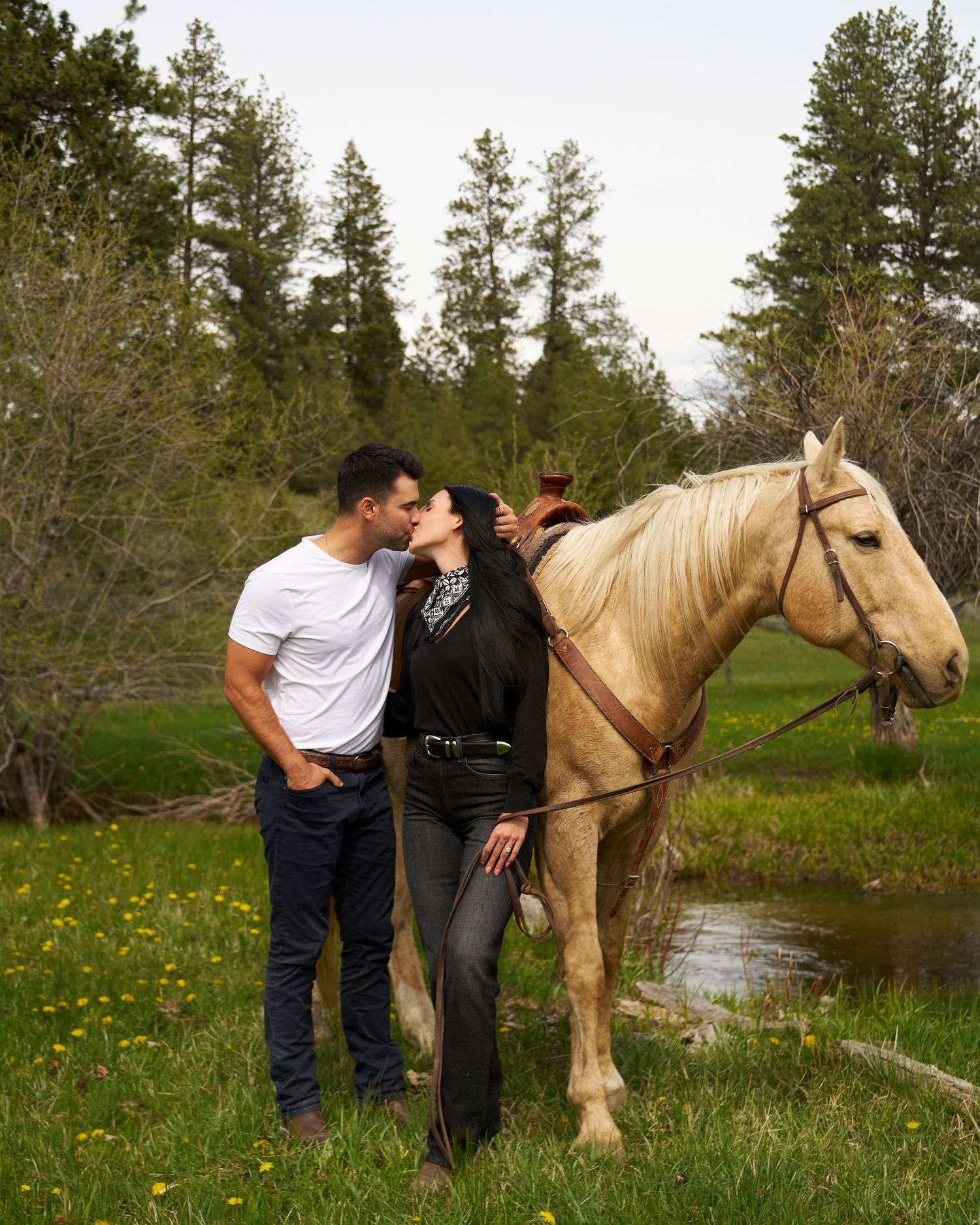 Couple sharing a kiss beside a palomino horse in a scenic woodland meadow.