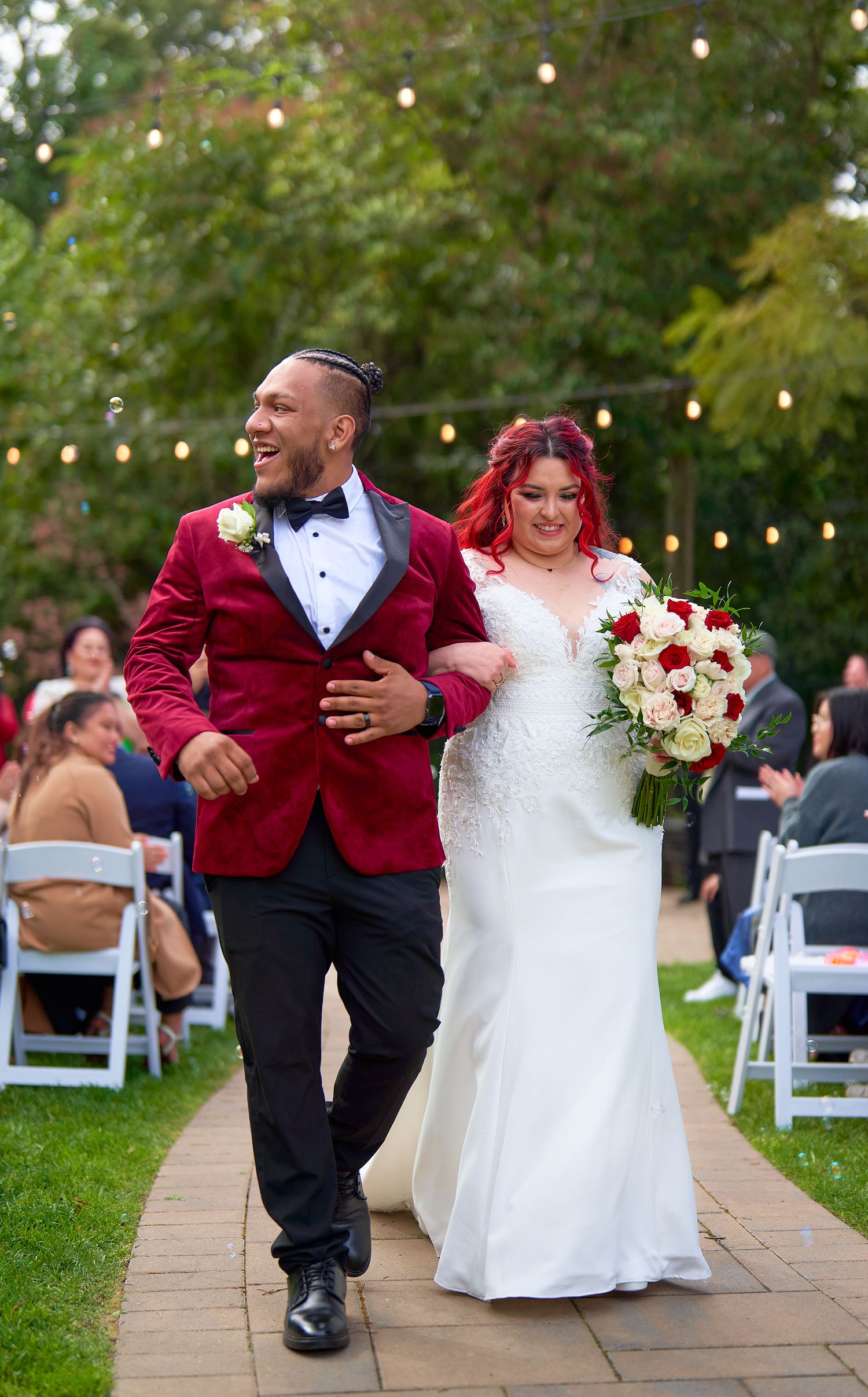 Joyful newlyweds walk down aisle, groom in red velvet blazer, bride holding red bouquet.
