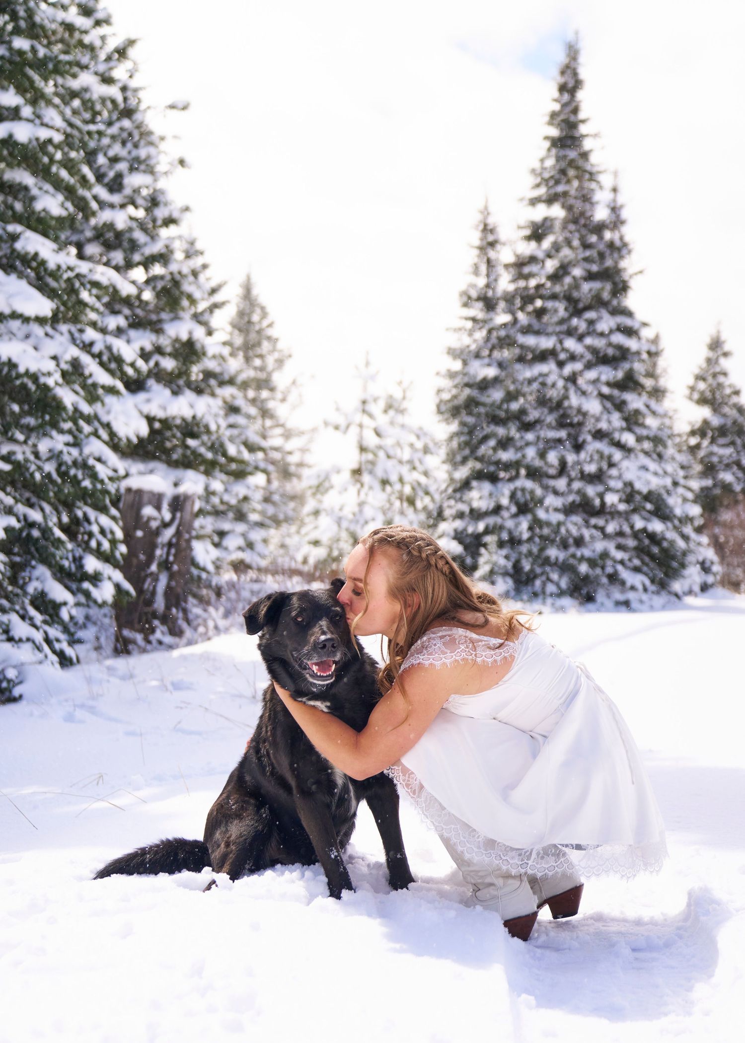 Bride in white lace dress kissing her black Labrador dog in a snowy winter forest setting.