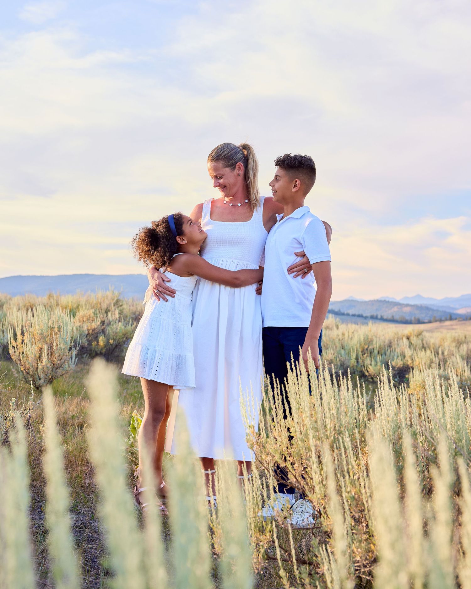 A mother in a white dress shares a joyful moment with her two children outdoors.