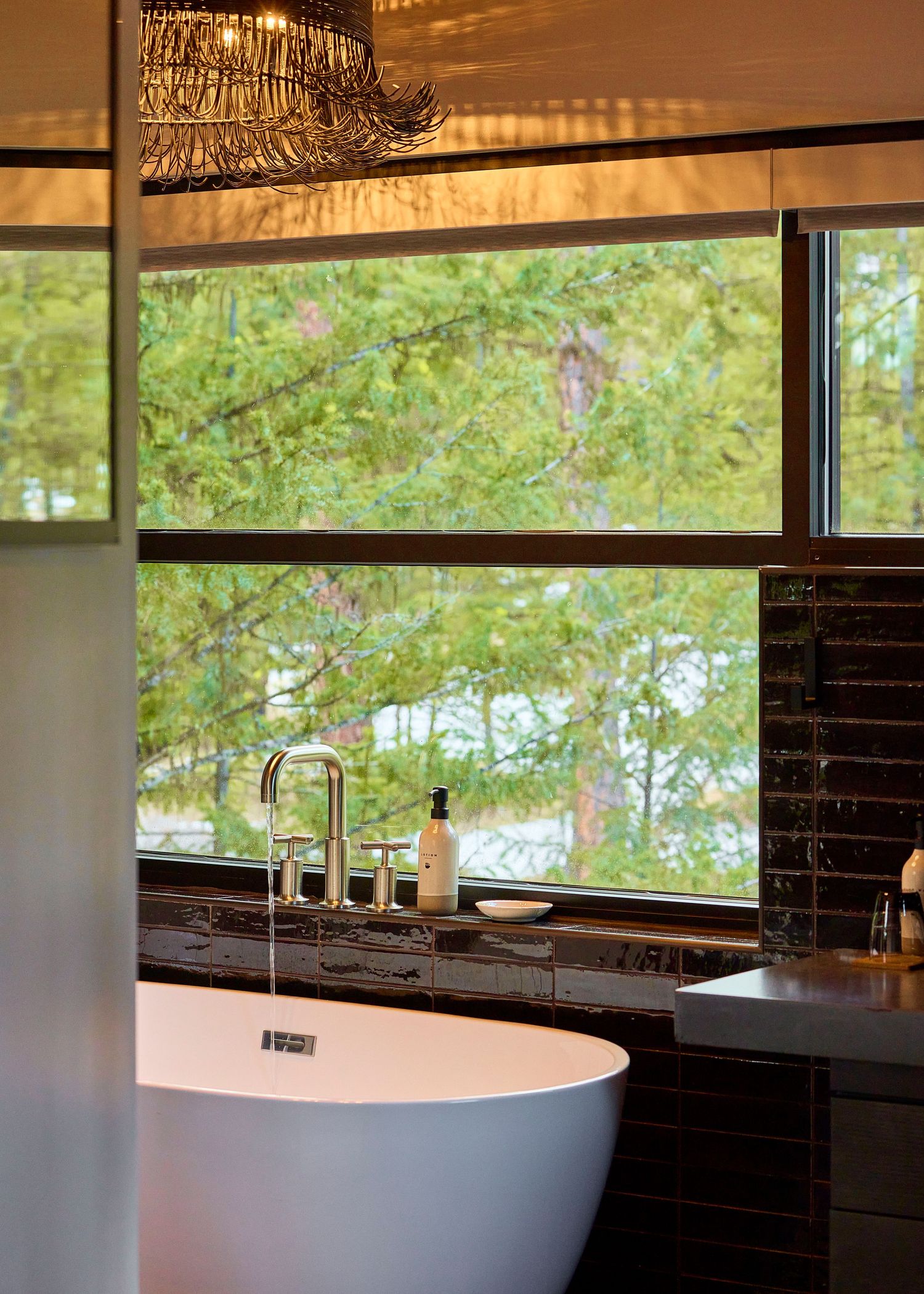 A freestanding white soaking tub beside a large window overlooking lush green pine trees in a luxury bathroom.
