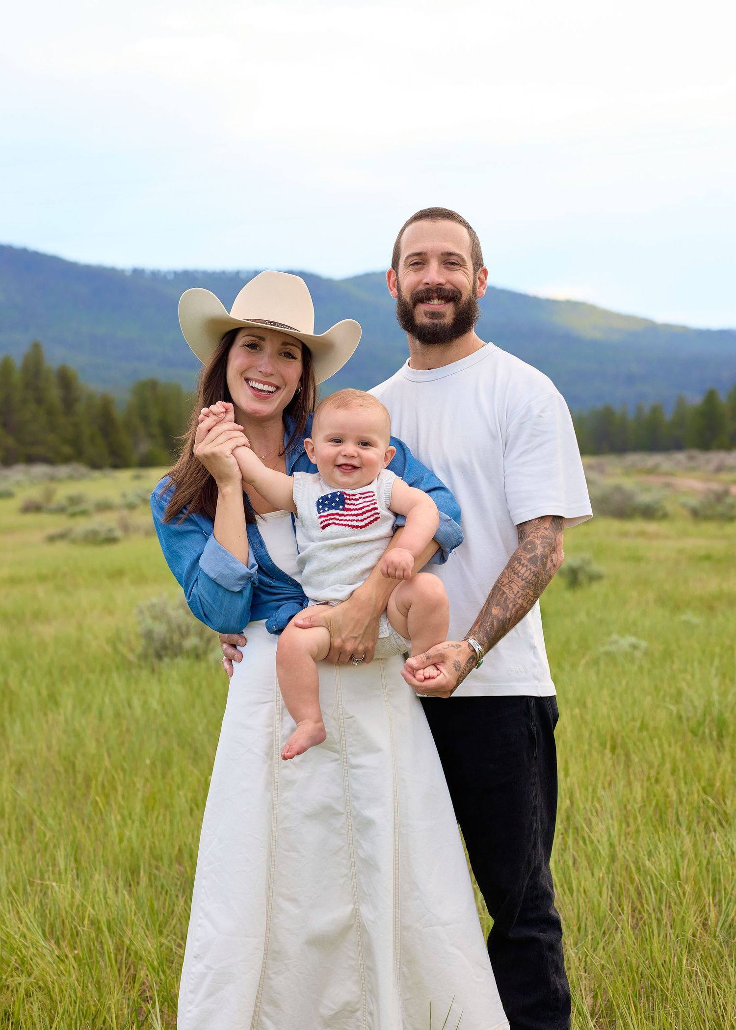Couple holding a smiling baby in an American flag onesie in a green mountain meadow.